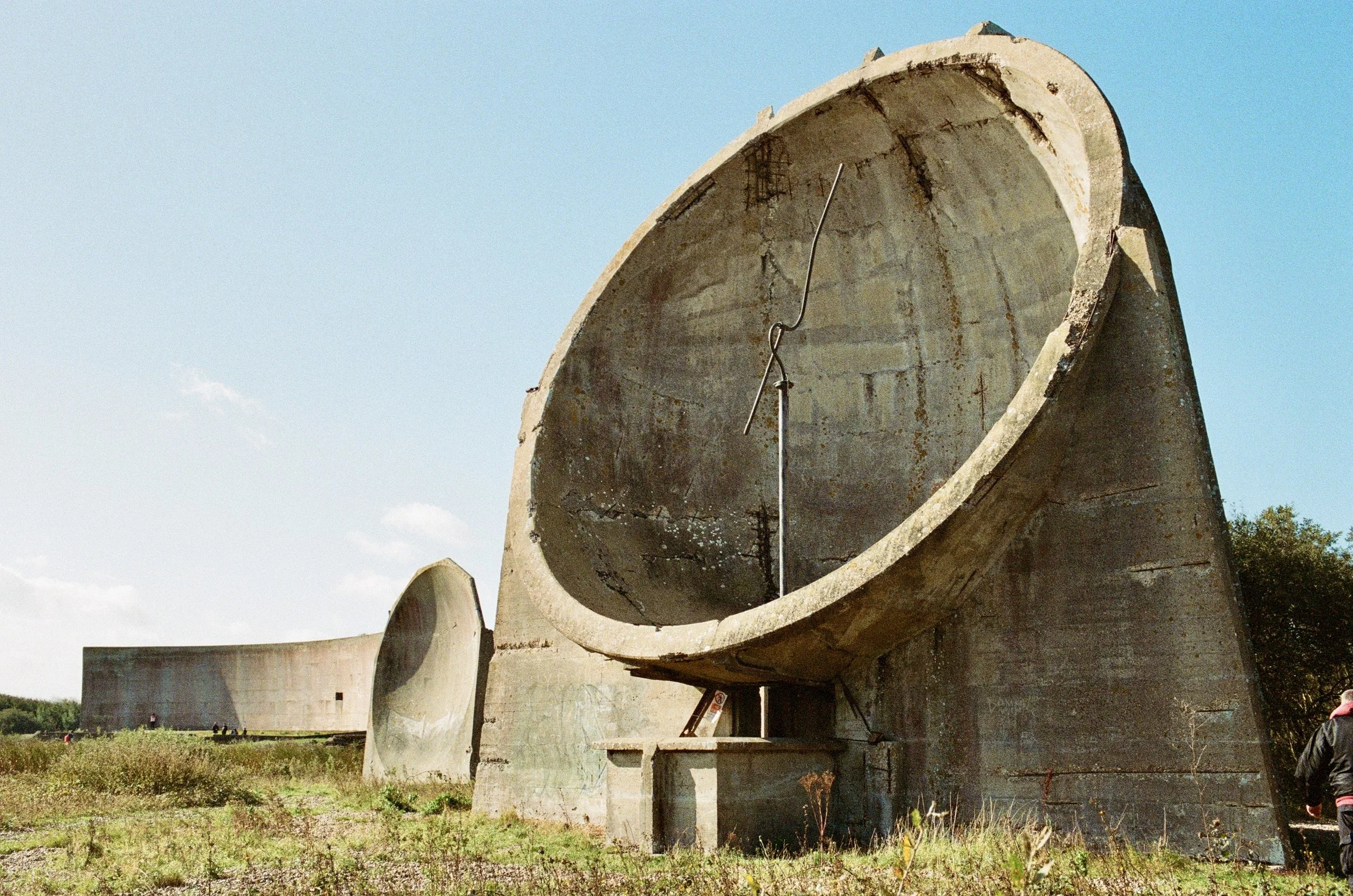 Large concrete structure resembling a rocket silo or underground storage tank with a curved opening and supporting legs, situated outdoors with a partly cloudy sky and trees in the background.