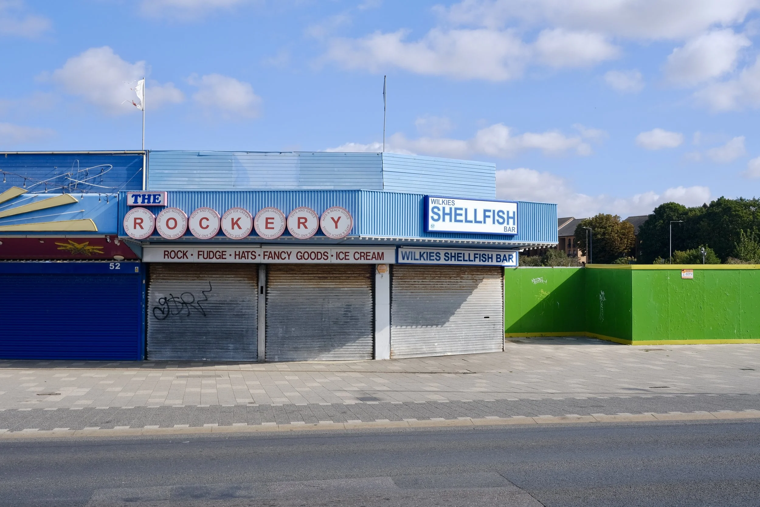 Closed storefront with signs reading 'The Rocket,' 'Wilkies Shellfish Bar,' and advertisements for rock, fudge, hats, fancy goods, and ice cream. Colorful panels and graffiti on shutters, with a green wall on the right and a clear blue sky above.