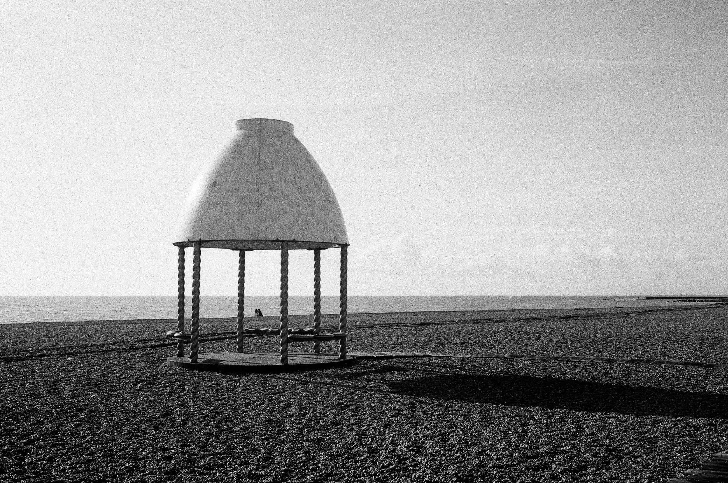 A black-and-white photo of a swing set on a beach with the ocean in the background. The swing set has a covered top and twisted rope supports, and there are two people sitting on the sand near the water.