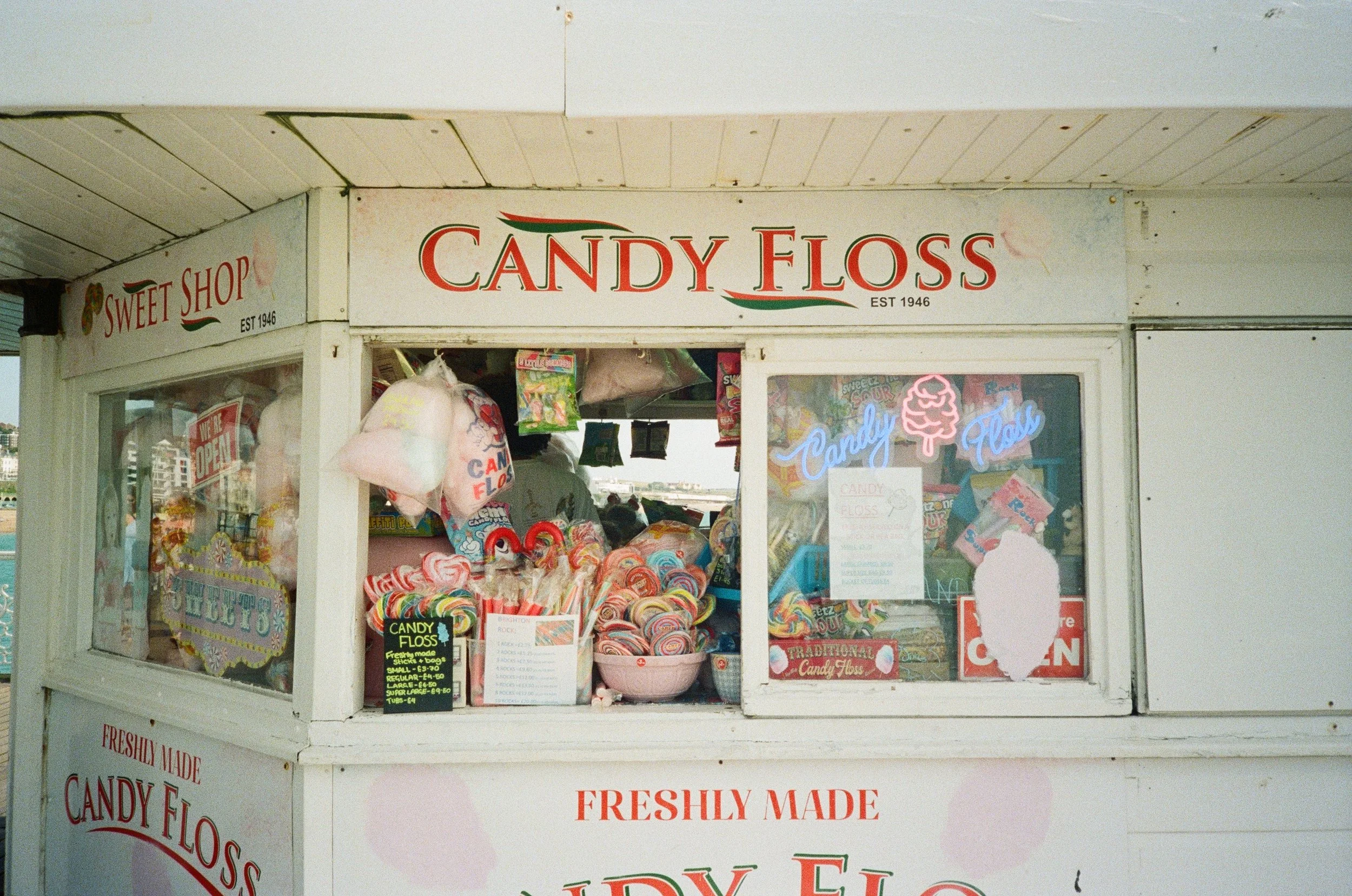 A candy floss stand with colorful candy floss on display, signage indicating the store's name 'Candy Floss' and establishment year 1946, along with a neon sign that says 'Candy Floss' and a pink cotton candy icon.