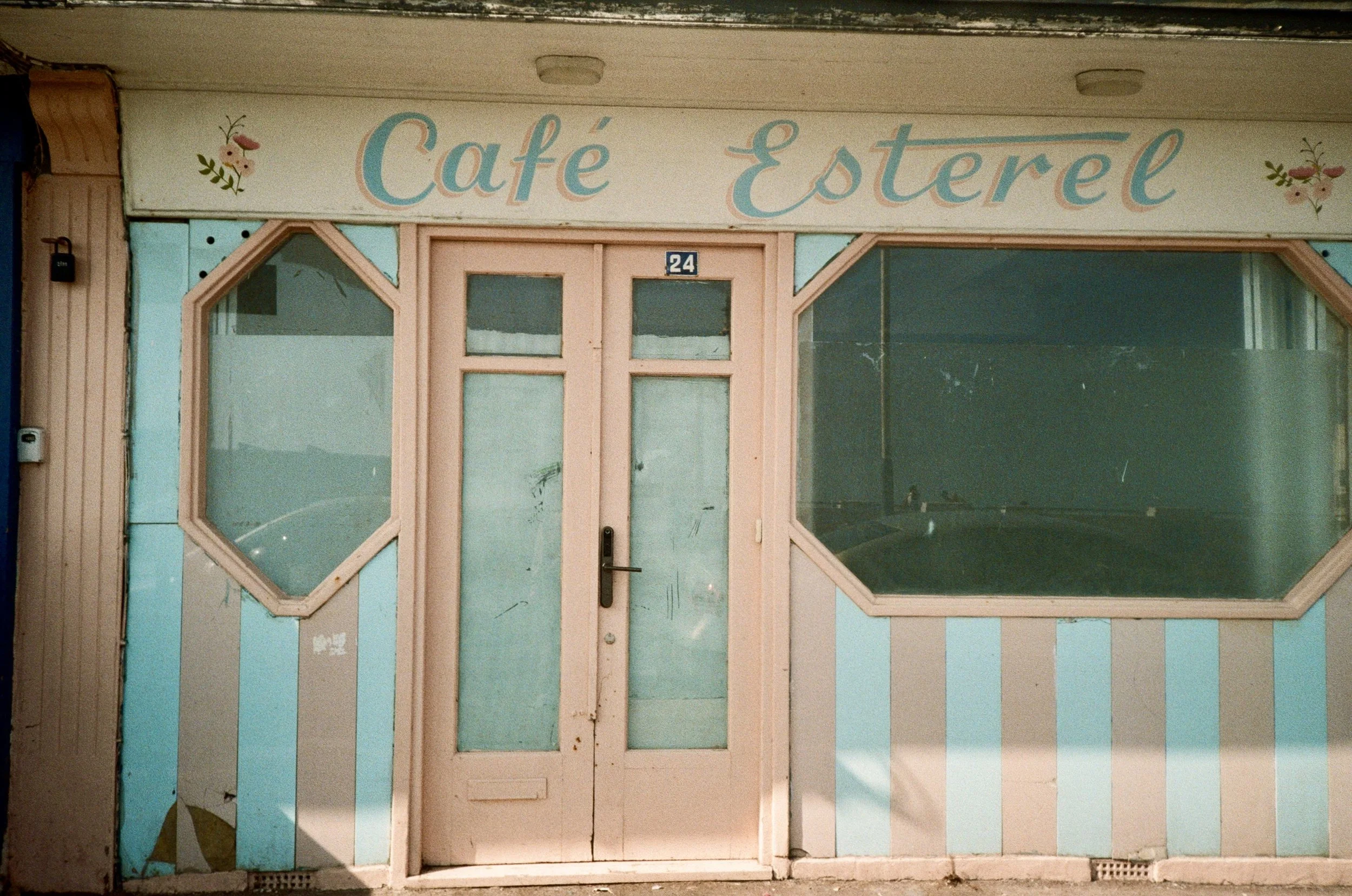 Exterior of a pastel-colored cafe with a sign reading 'Café Estéer' above the door and large windows on either side, with the building number 24 displayed above the door.