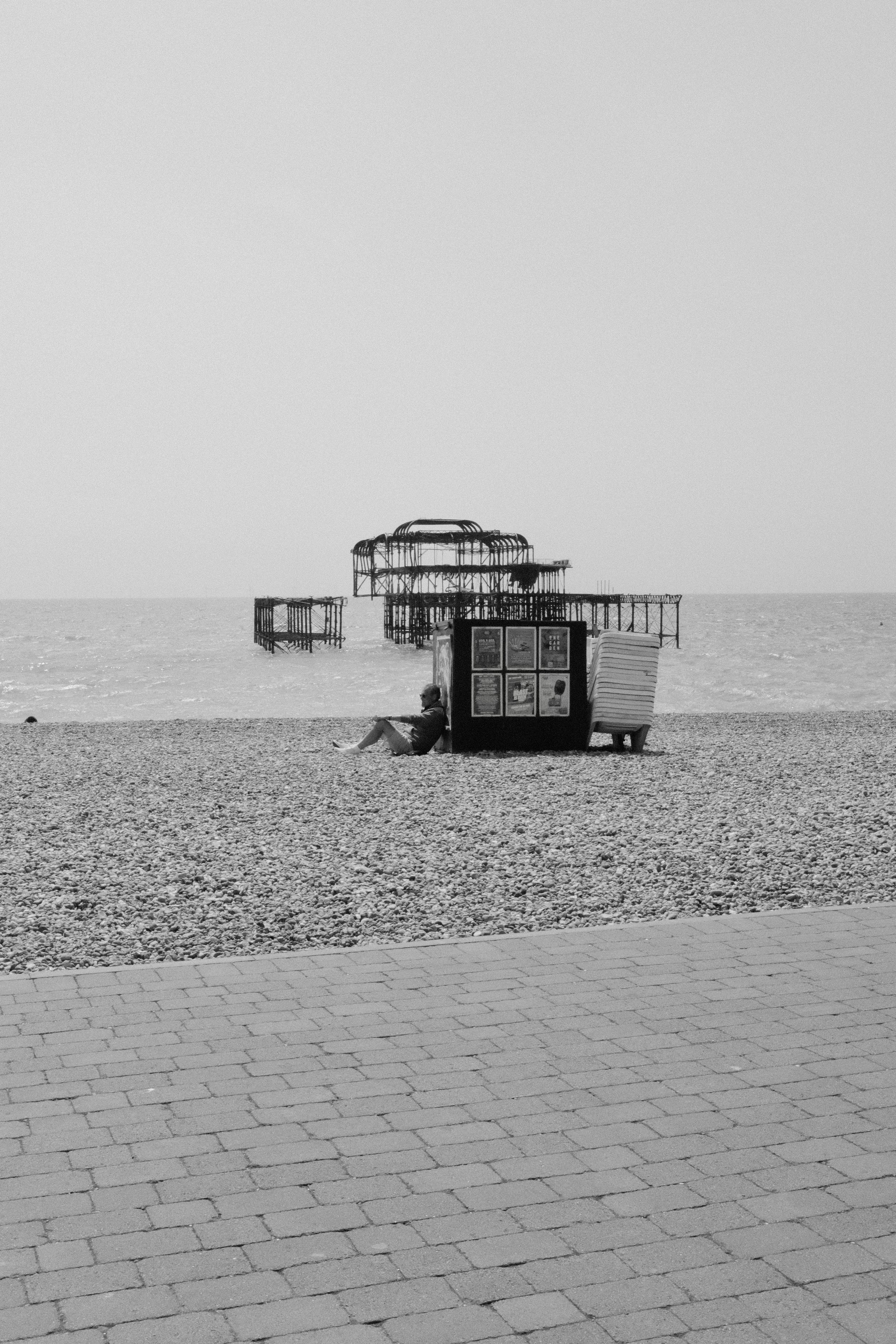 A person sitting on a bench near the beach with a large, dilapidated pier structure in the background.