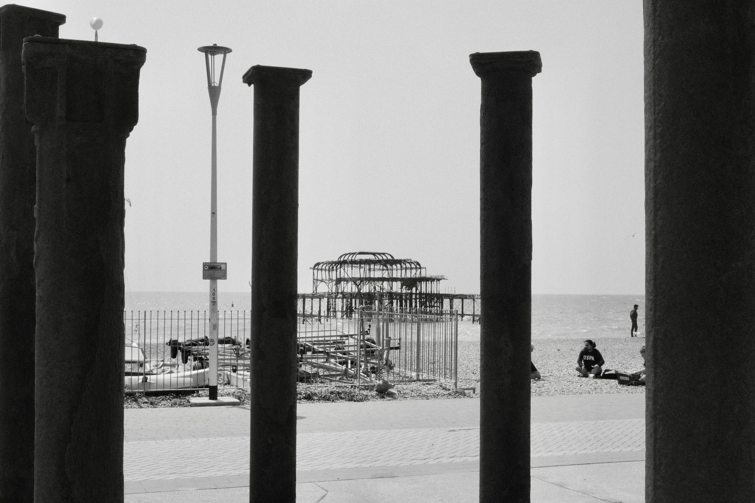 View of a beach scene through four large stone pillars. In the background, there is an outdoor structure near the water, a person wearing a U.S. Postal Service jacket, and a person standing near the shoreline.