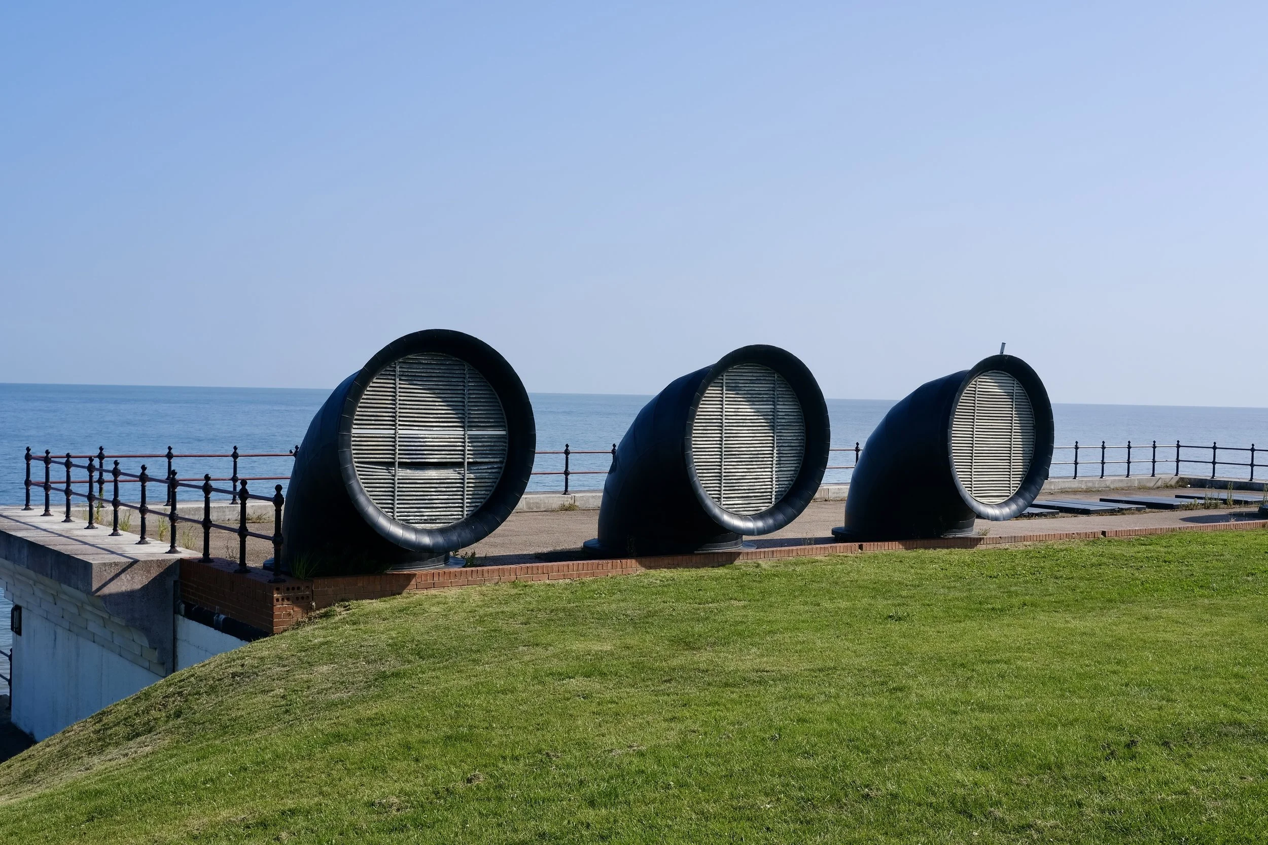 Three large black pipes with metal grates on an oceanfront promenade, with a blue sky and calm sea in the background.
