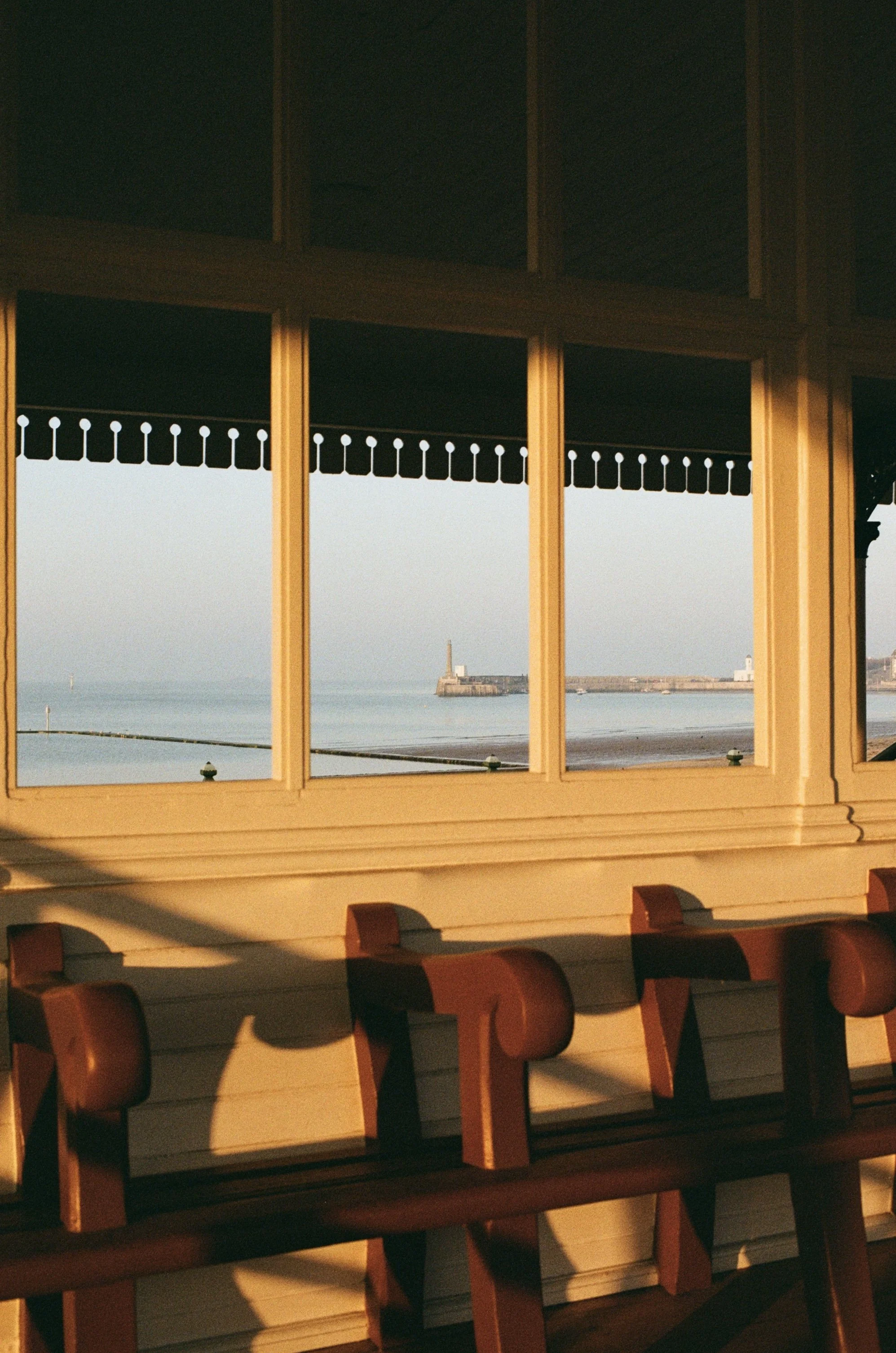 View of a beach scene through a window with cream-colored frame and brown trim. The scene includes a small boat on the water, a breakwater with a lighthouse or small building, and a clear sky.