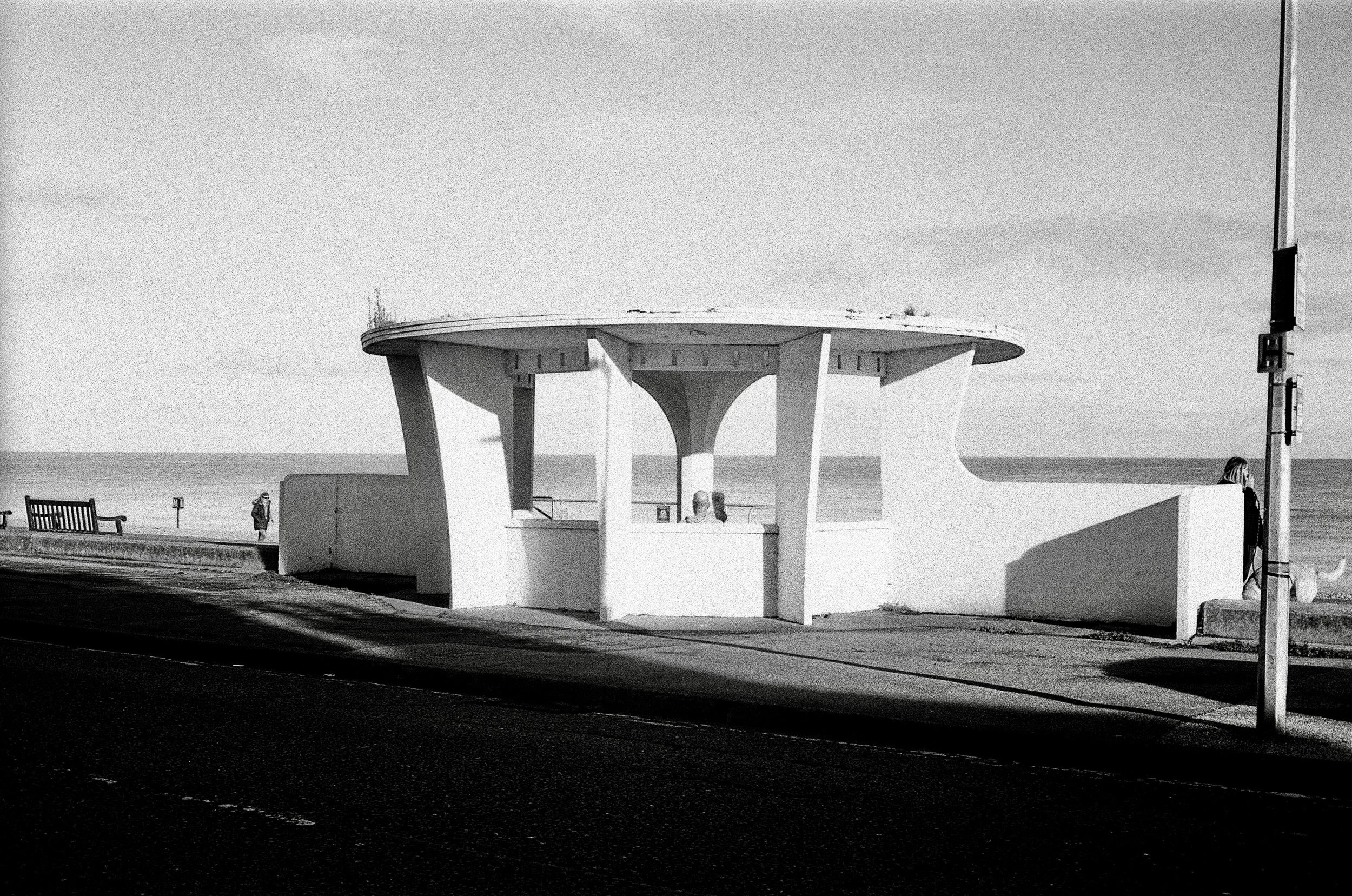 A black and white photo of a circular structure with supporting columns, located near the beach. There are a few people in the scene, some sitting and walking, with the ocean visible in the background. A flagpole is seen on the right side of the imag