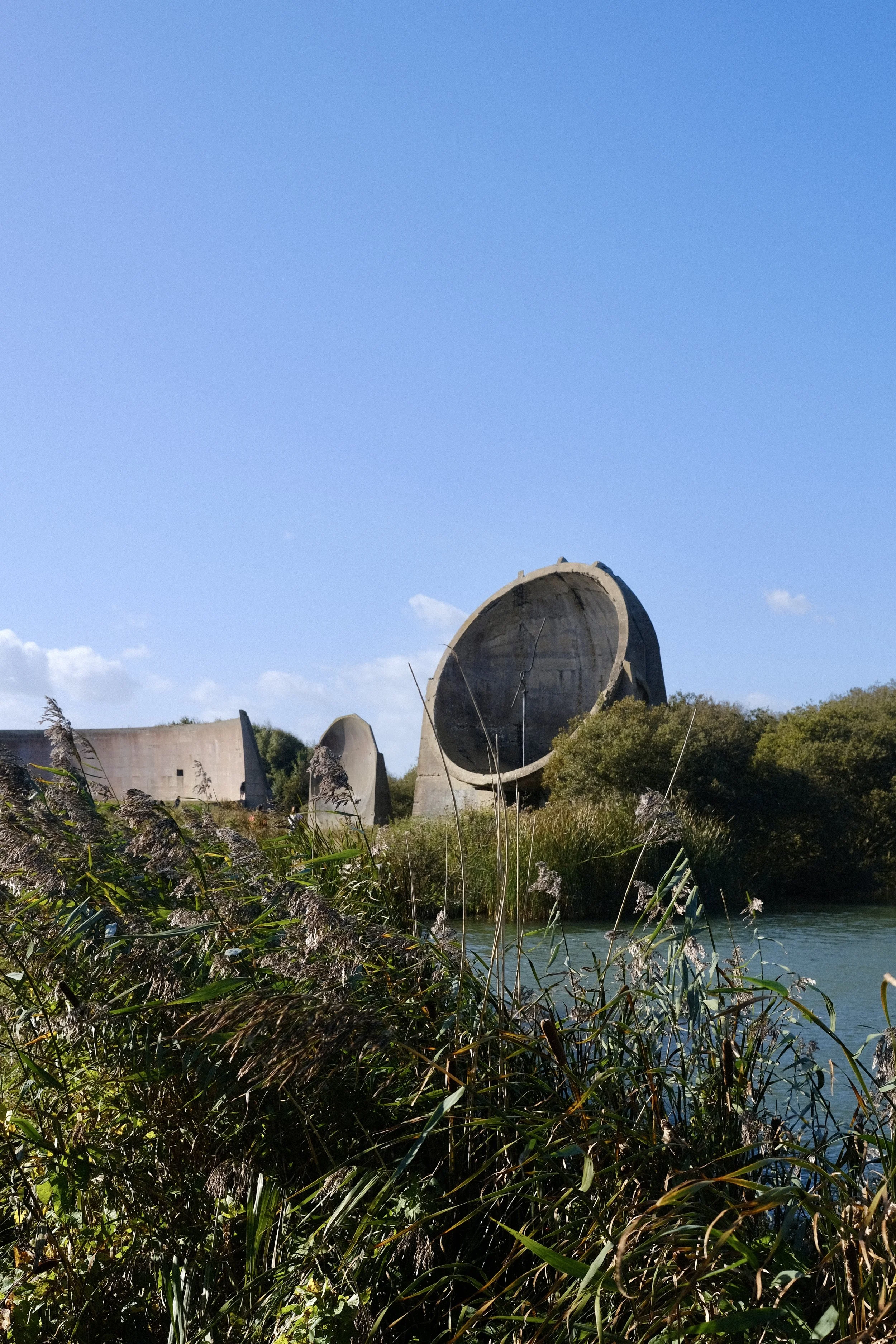 Large concrete structures resembling the remains of old industrial turbines or water treatment facilities are situated near a river, surrounded by dense green vegetation, with a clear blue sky overhead.