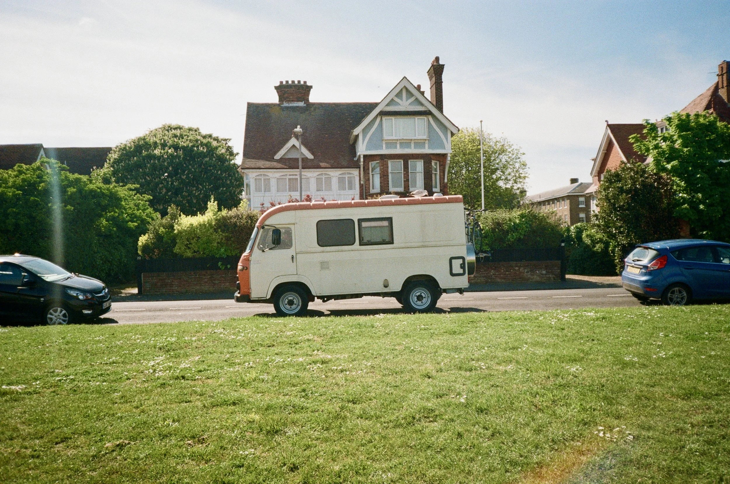 Residential street scene with a vintage camper van parked among other cars, in front of houses surrounded by greenery and trees.