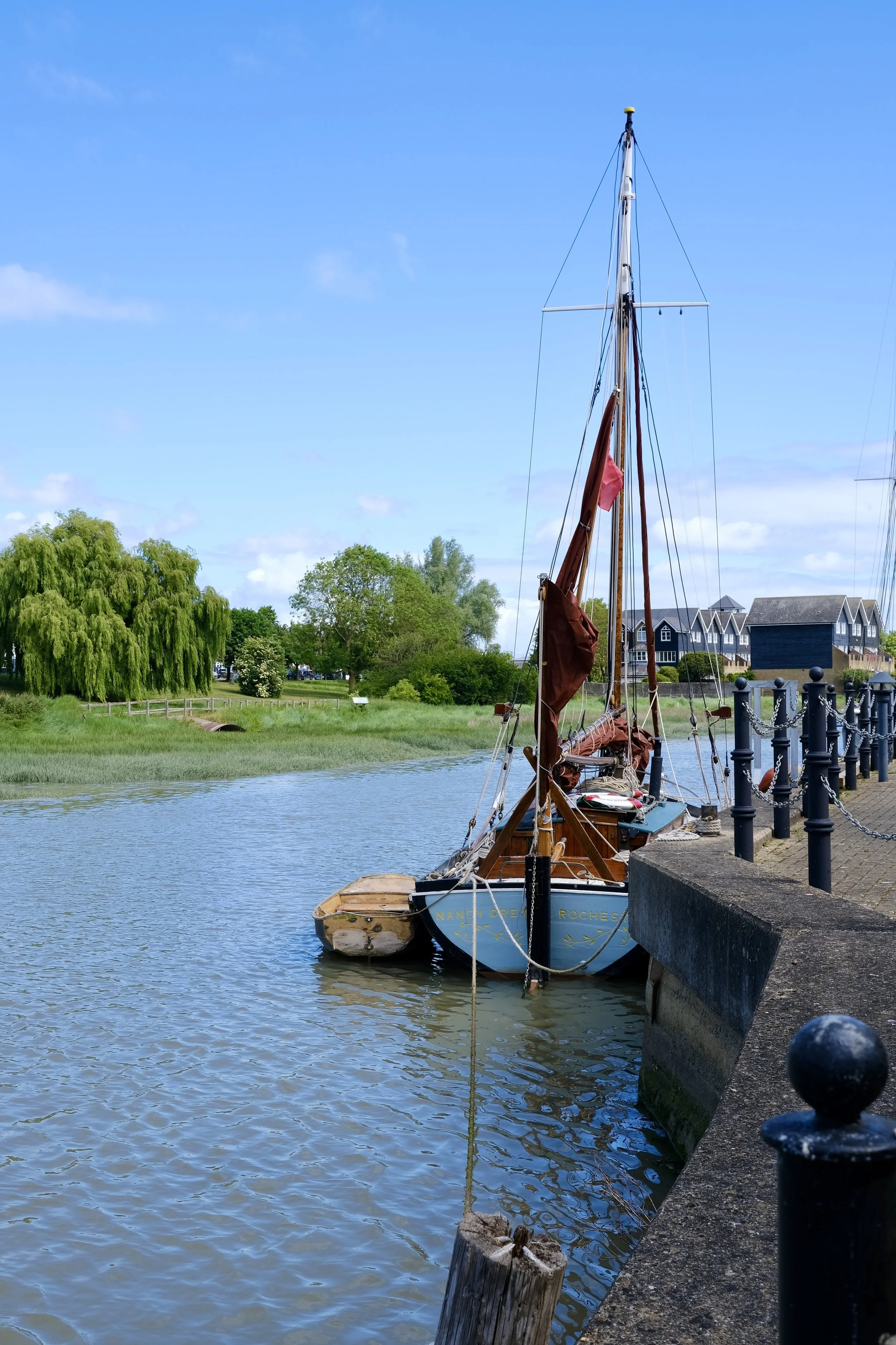 A sailboat docked along a riverbank with lush green trees and houses in the background, under a blue sky with scattered clouds.