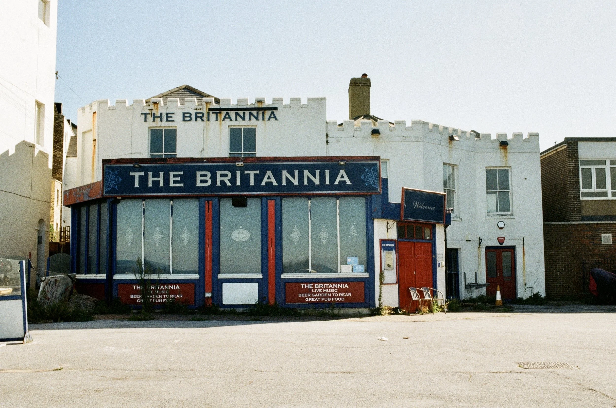 A building with signage reading 'The Britannia' and an outdoor area with chairs and signage promoting live music, a beer garden, and pub food.