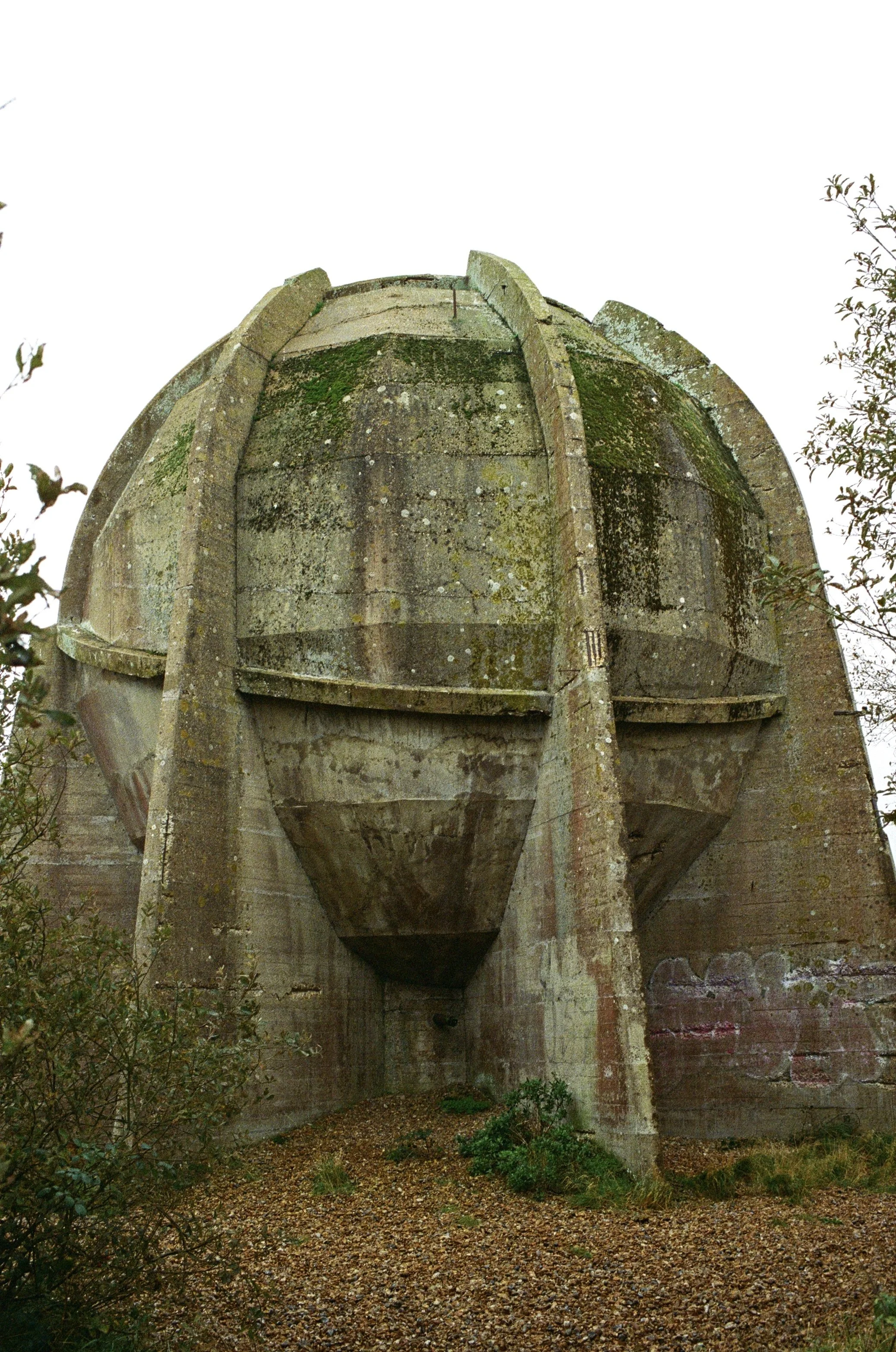 A large, ancient, weathered stone fountain with moss and lichen, surrounded by overgrown plants and trees, with a bright white sky in the background.