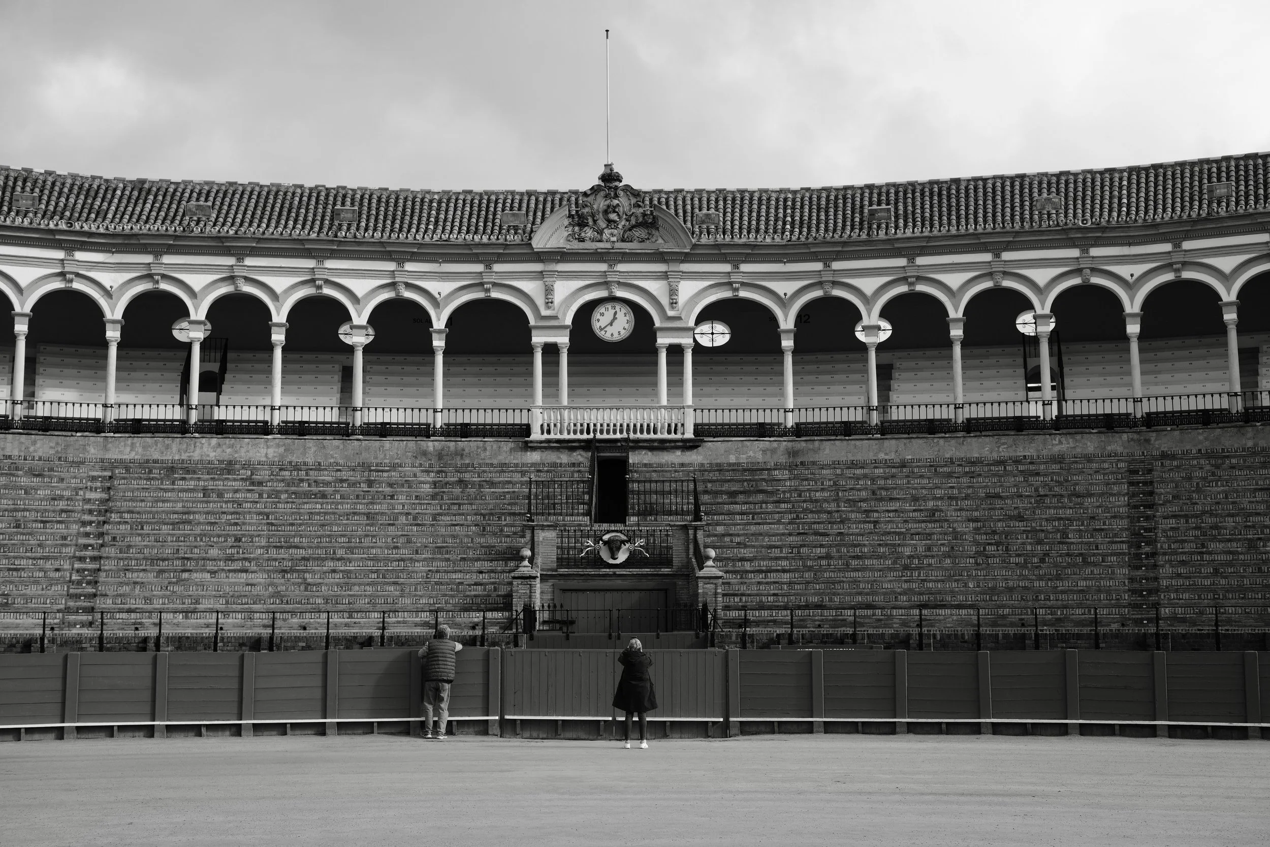 A black-and-white photo of an empty arena with tiered seating, a clock, and a decorative crest above the seating area. Three people are standing in front of a fence, facing the arena.