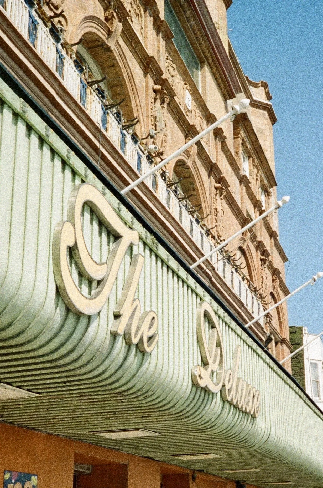 The photo shows the exterior of a building with an ornate upper facade featuring arched windows and decorative details. Below, there is a green, horizontally-striped awning with cursive lettering that reads 'The '. White streetlights are mounted on t