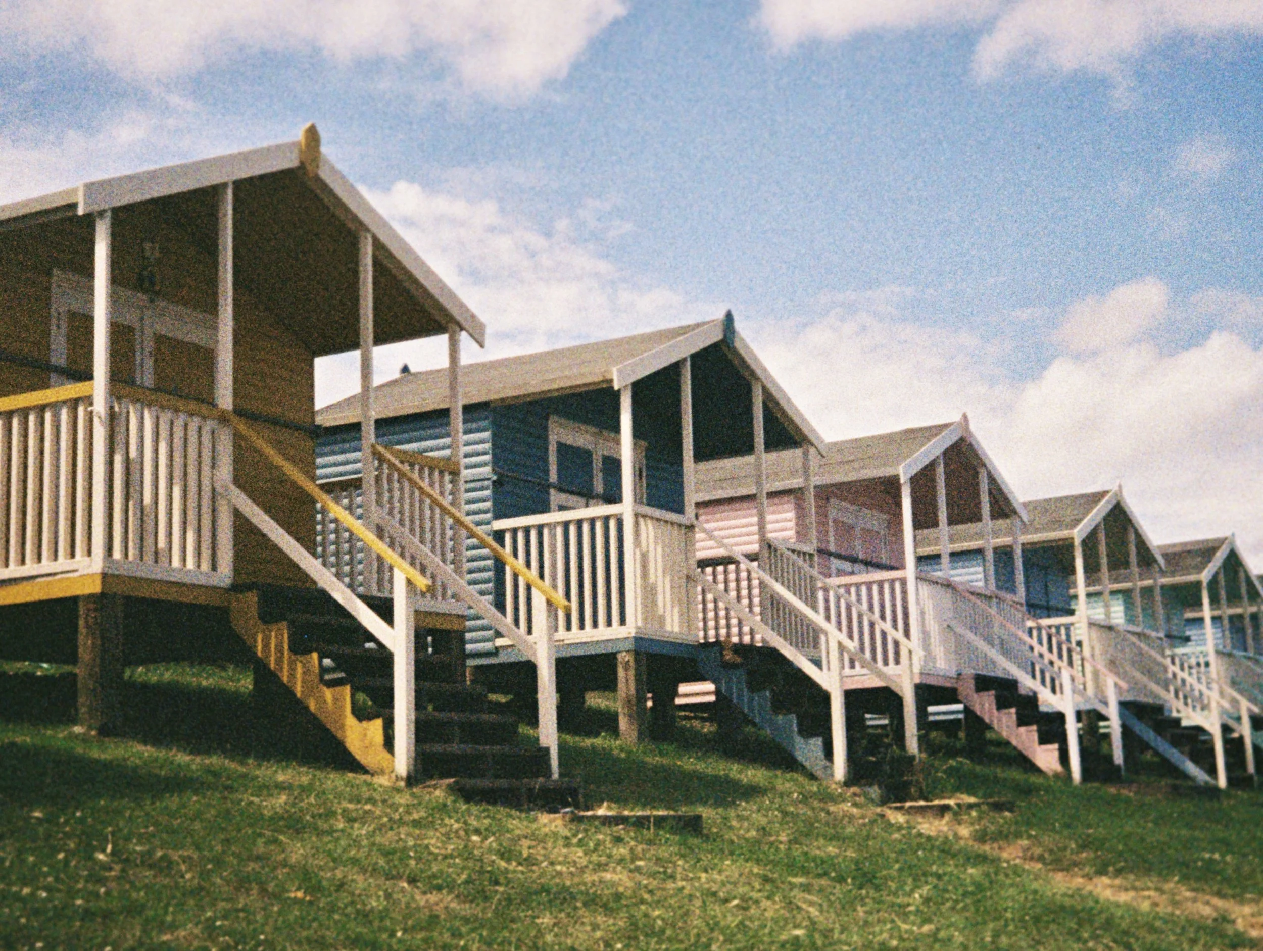 Colorful beach houses on stilts with stairs and decks, under a cloudy sky, on a grassy area.