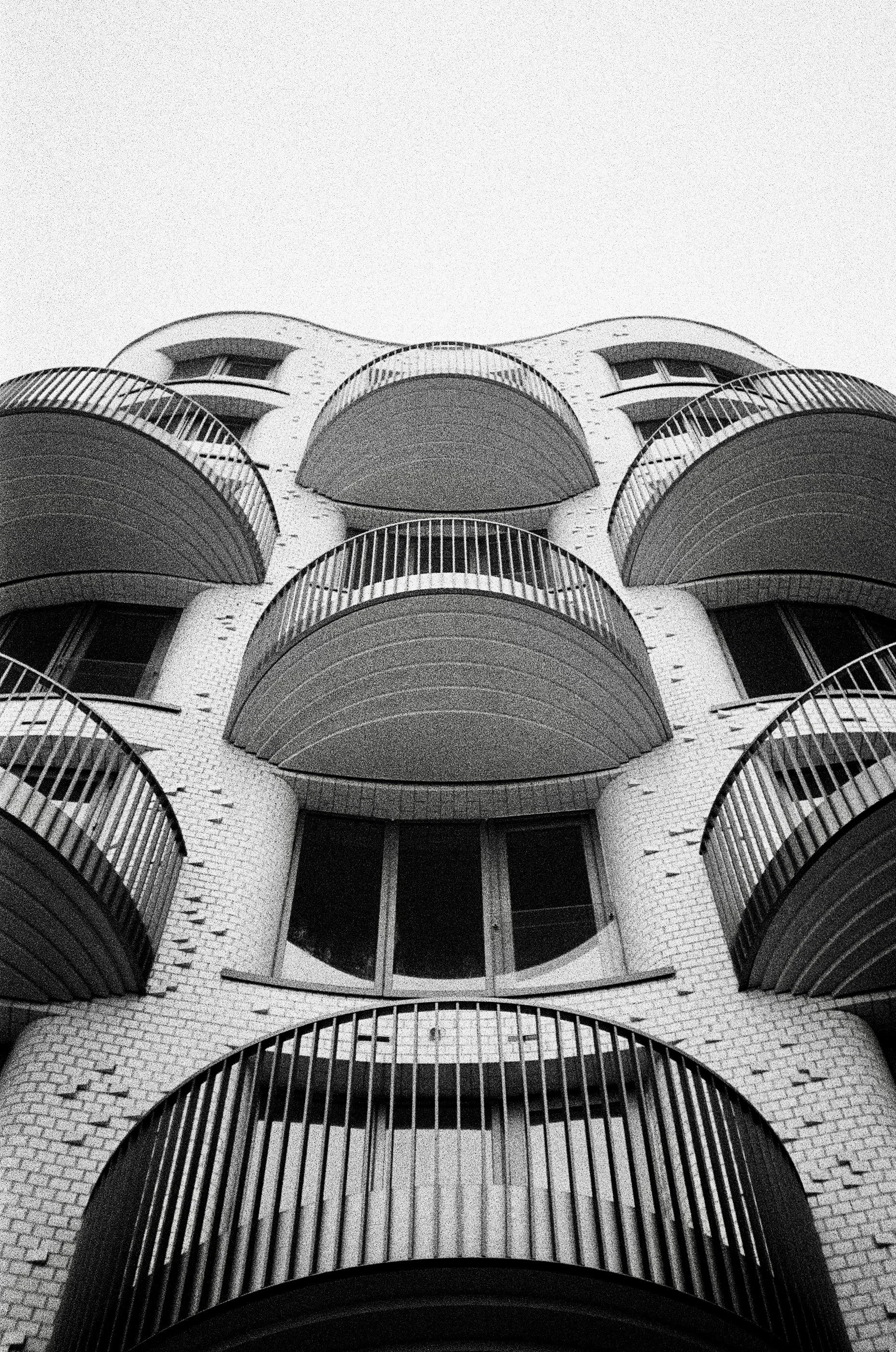 Low-angle view of a modern multi-story building with curved balconies and white brick exterior, black window frames, and metal railings, viewed from the ground looking up.