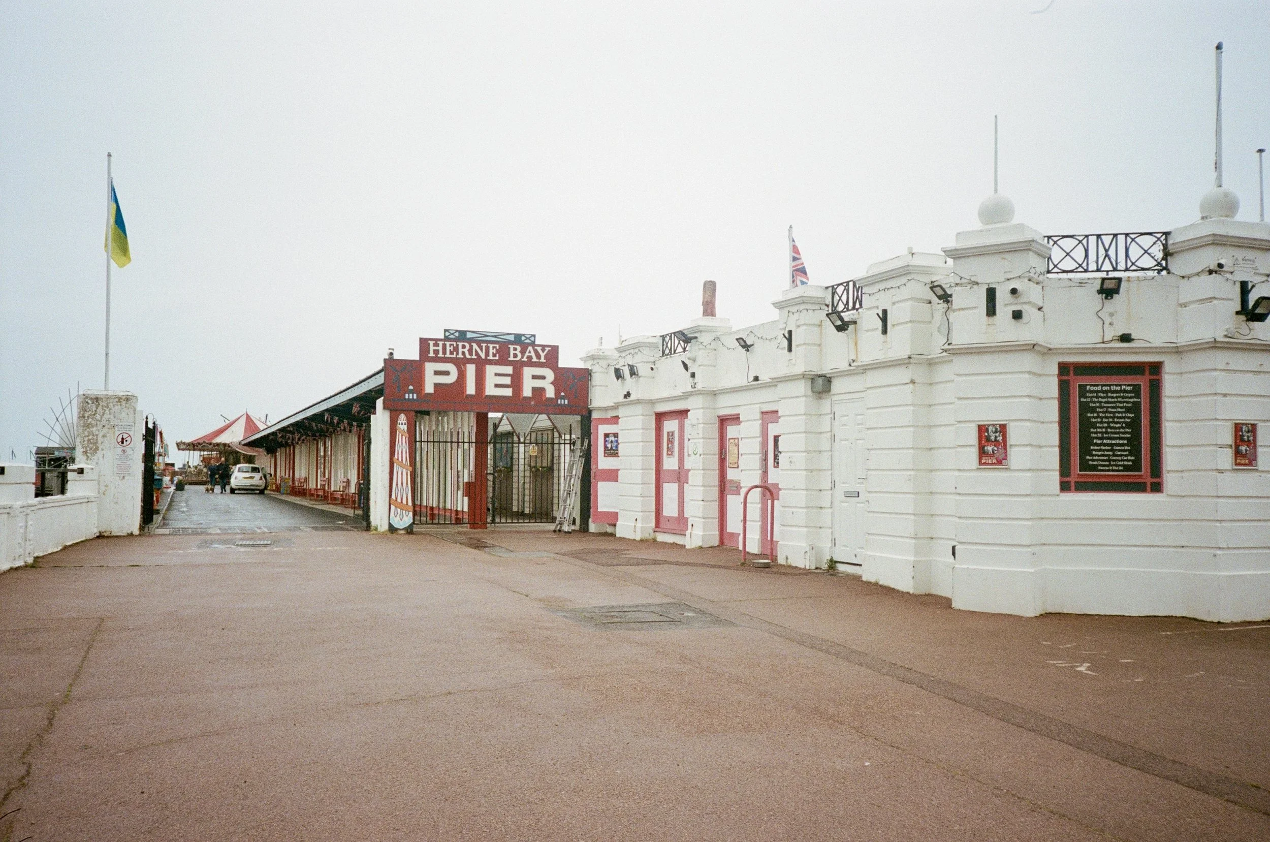 The image shows the entrance to Herne Bay Pier with flags, a sign, and a white building, under an overcast sky.