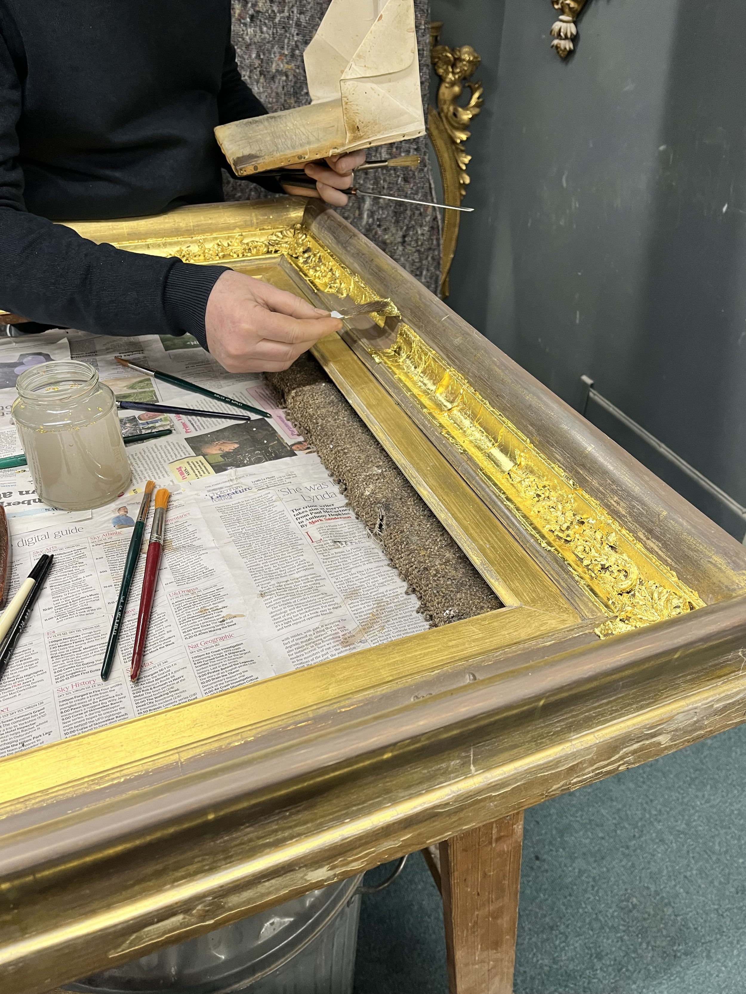 Person working on restoring or gilding a picture frame with a small paintbrush and a wooden stick, with newspapers spread on the table for protection, in an indoor setting with dark walls and ornate gold decorations in the background.