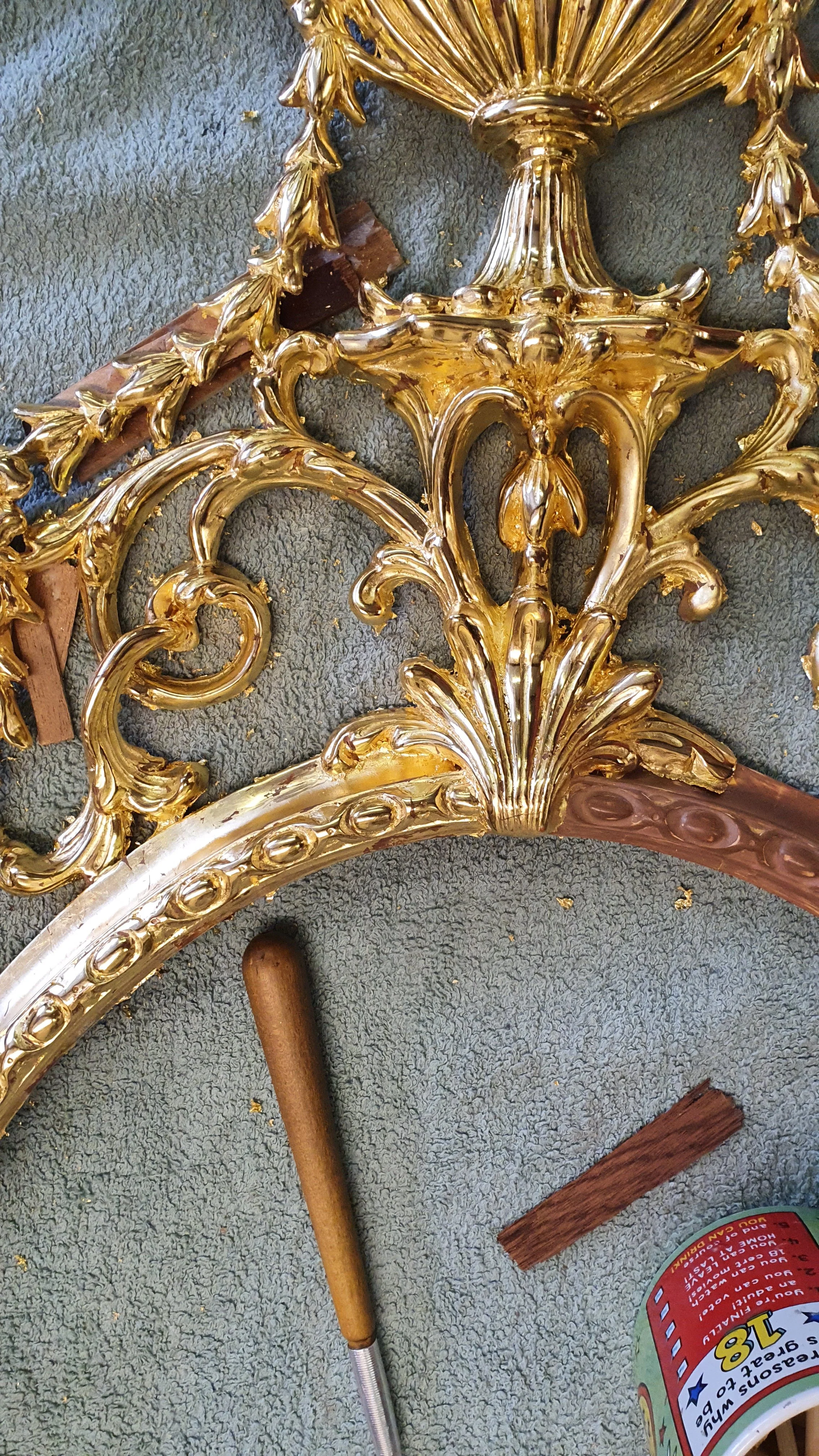 Close-up of a broken ornate gold-framed mirror or picture frame with intricate decorative details, laying on a textured gray surface with various small objects and debris around.