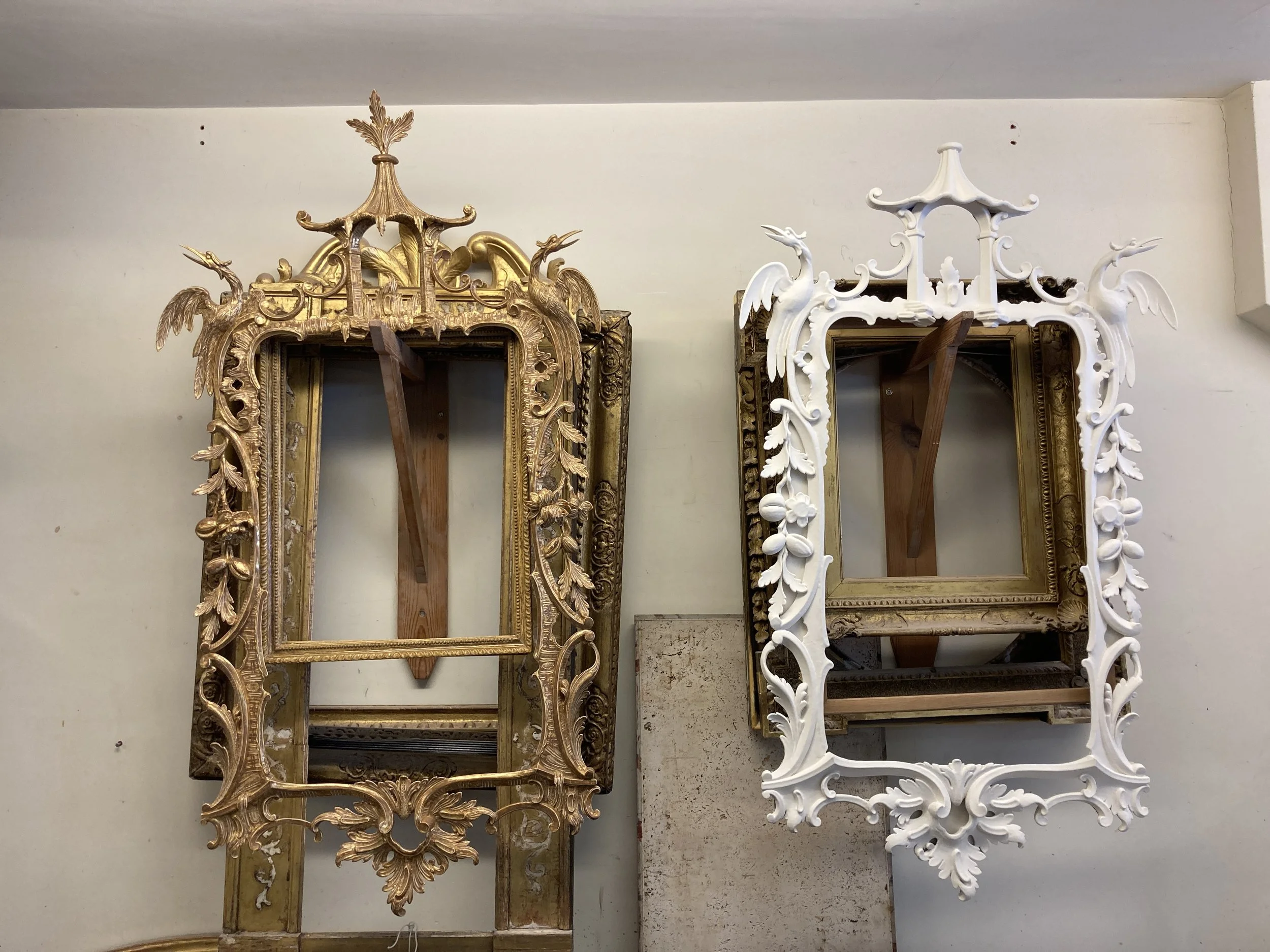 Two ornate, decorative picture frames, one gold and one white, positioned against a plain wall.