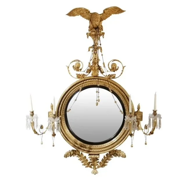An ornate gold-framed mirror with a circular reflective surface, flanked by two crystal candle holders with candles, topped with a decorative eagle sculpture.
