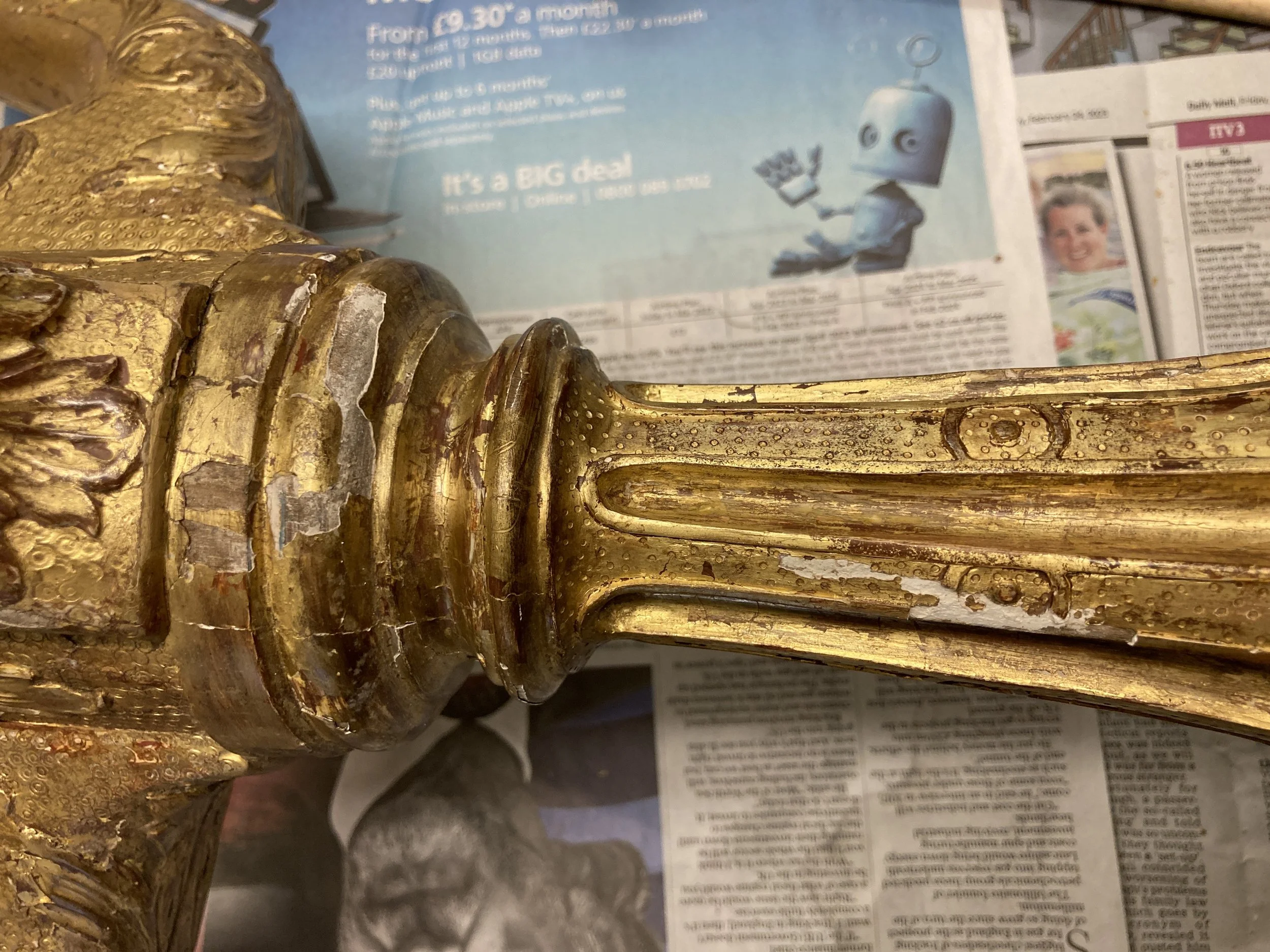 Close-up of a gold ornate decorative object, possibly a part of a frame or furniture, with a background showing newspaper advertisements.