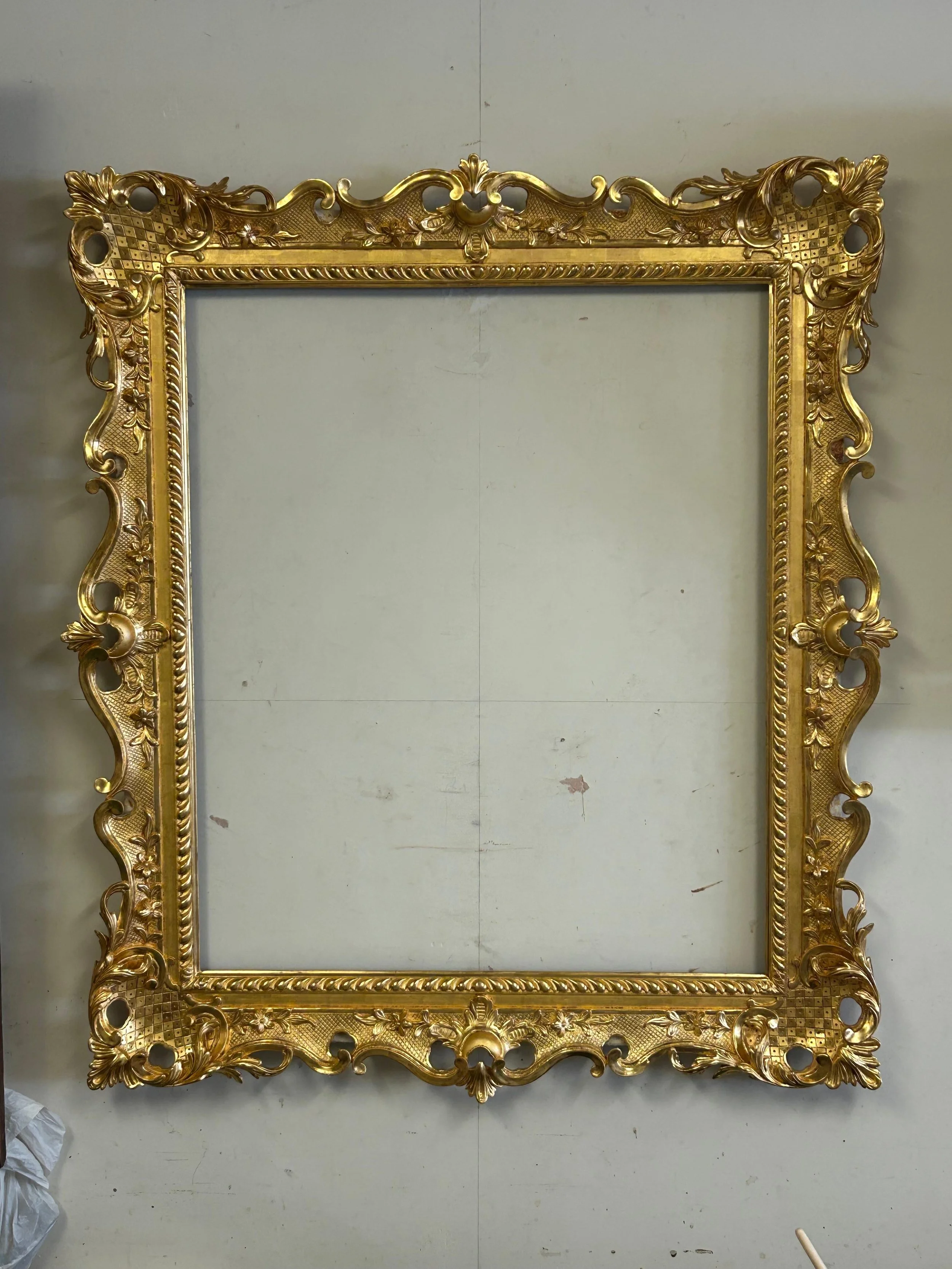 A large ornate gold picture frame with intricate scrollwork and floral patterns, resting against a plain gray wall.