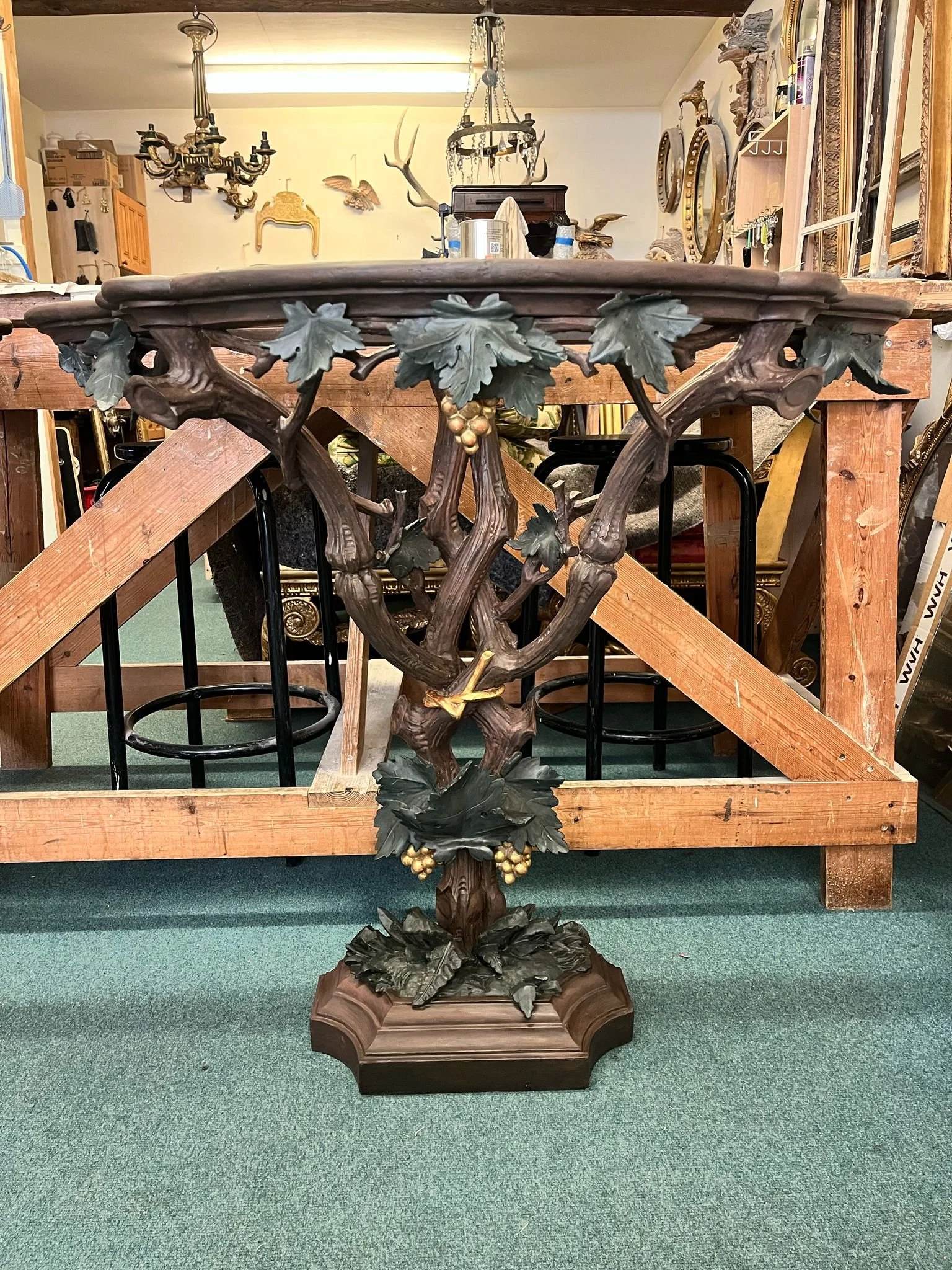 A decorative wooden pedestal table with a leaf and grapevine motif, featuring grape clusters and leaves, stands in front of a wooden railing and a workshop or antique store interior with various frames and decorations on the wall.