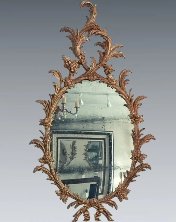 Ornate vintage oval mirror with a decorative gold frame, reflecting a room with paintings, a chandelier, and a ceiling light.