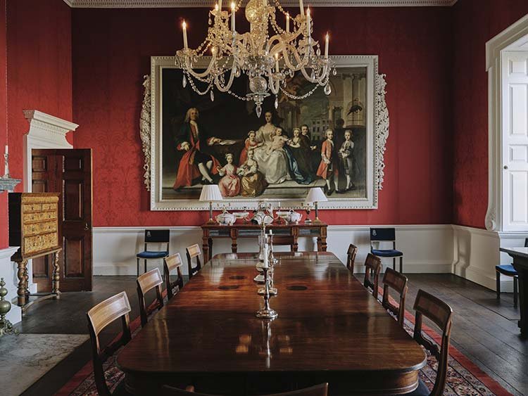 A formal dining room with a large wooden table and eight chairs, a chandelier hanging from the ceiling, a large historical painting on a red wall with white trim, and a buffet with lamps and decorative items.