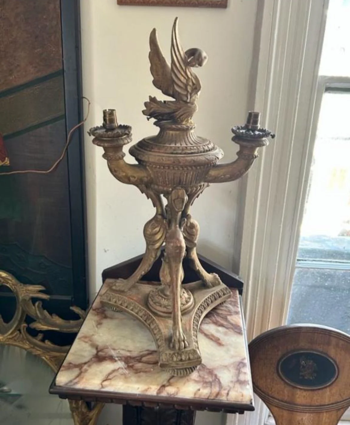 An ornate brass or bronze candelabrum with a swan figure at the top, featuring two arms for candles and a decorative base, placed on a marble-topped table near a window.