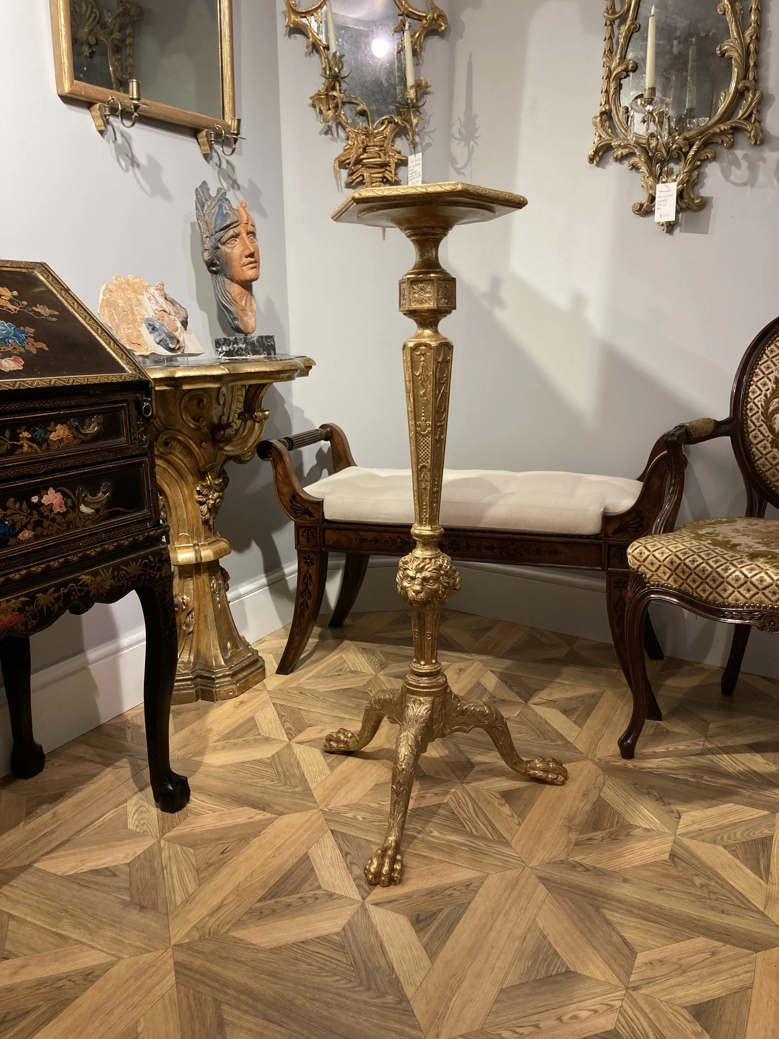 Antique gold-colored pedestal with lion paw feet in a room with ornate mirrors, wooden furniture with floral inlay, and a carved wooden chair with patterned upholstery.