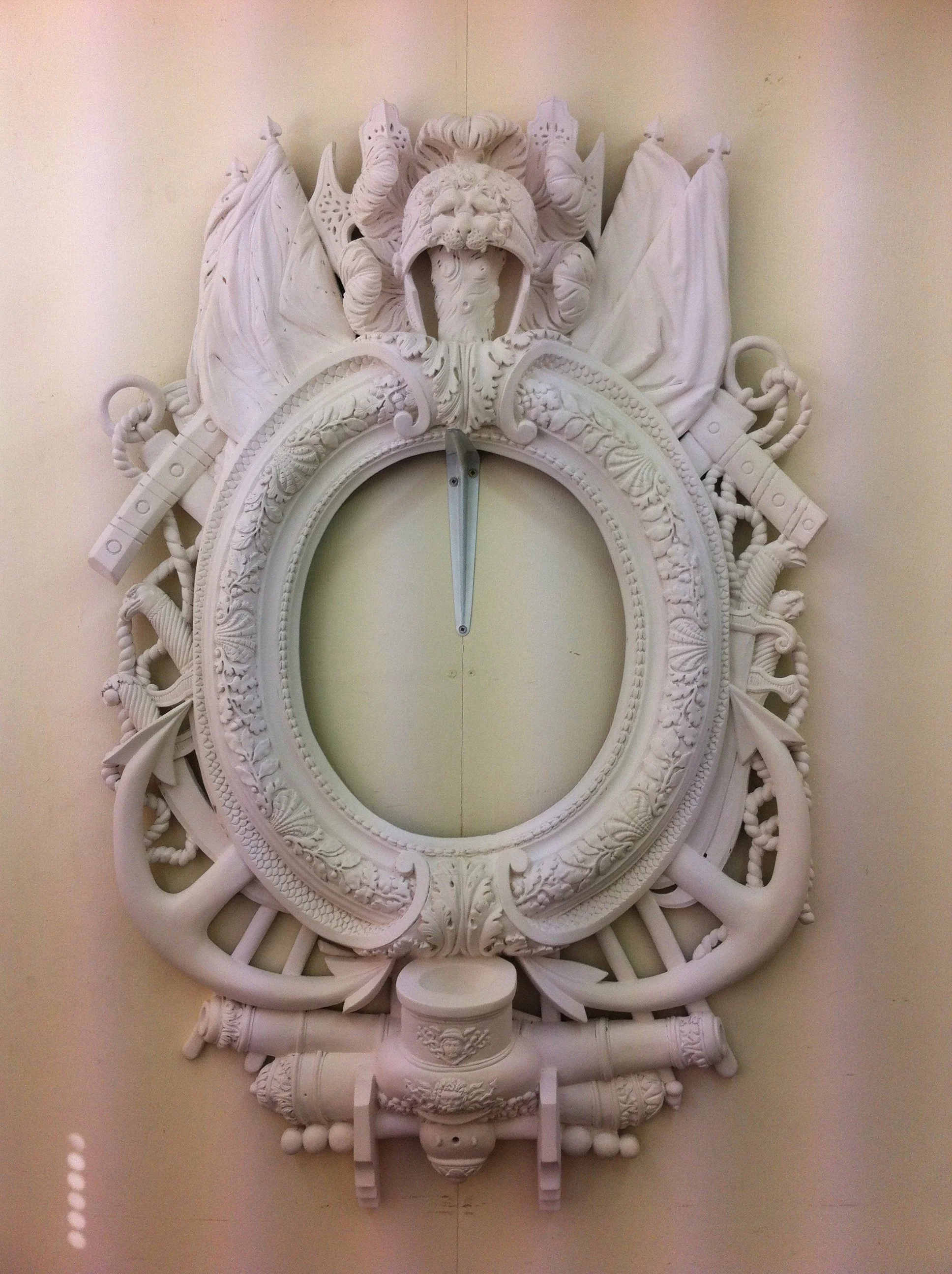 A white, ornate, decorative clock with a lion's head at the top and intricate carvings, including flags and scrolls, on the frame.