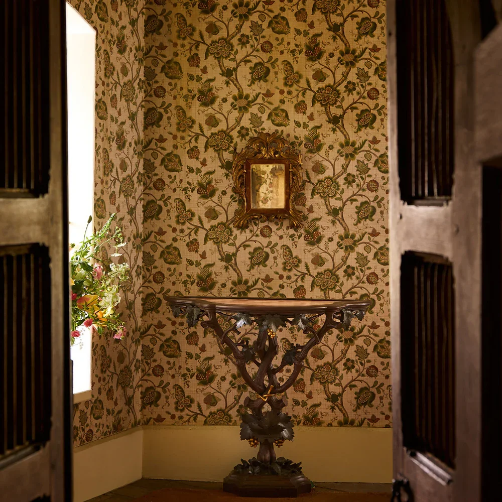 A small, ornate wooden table with intricate carvings, set against a floral patterned wallpaper wall, with a small framed mirror above it and a window with flowers on the left side.