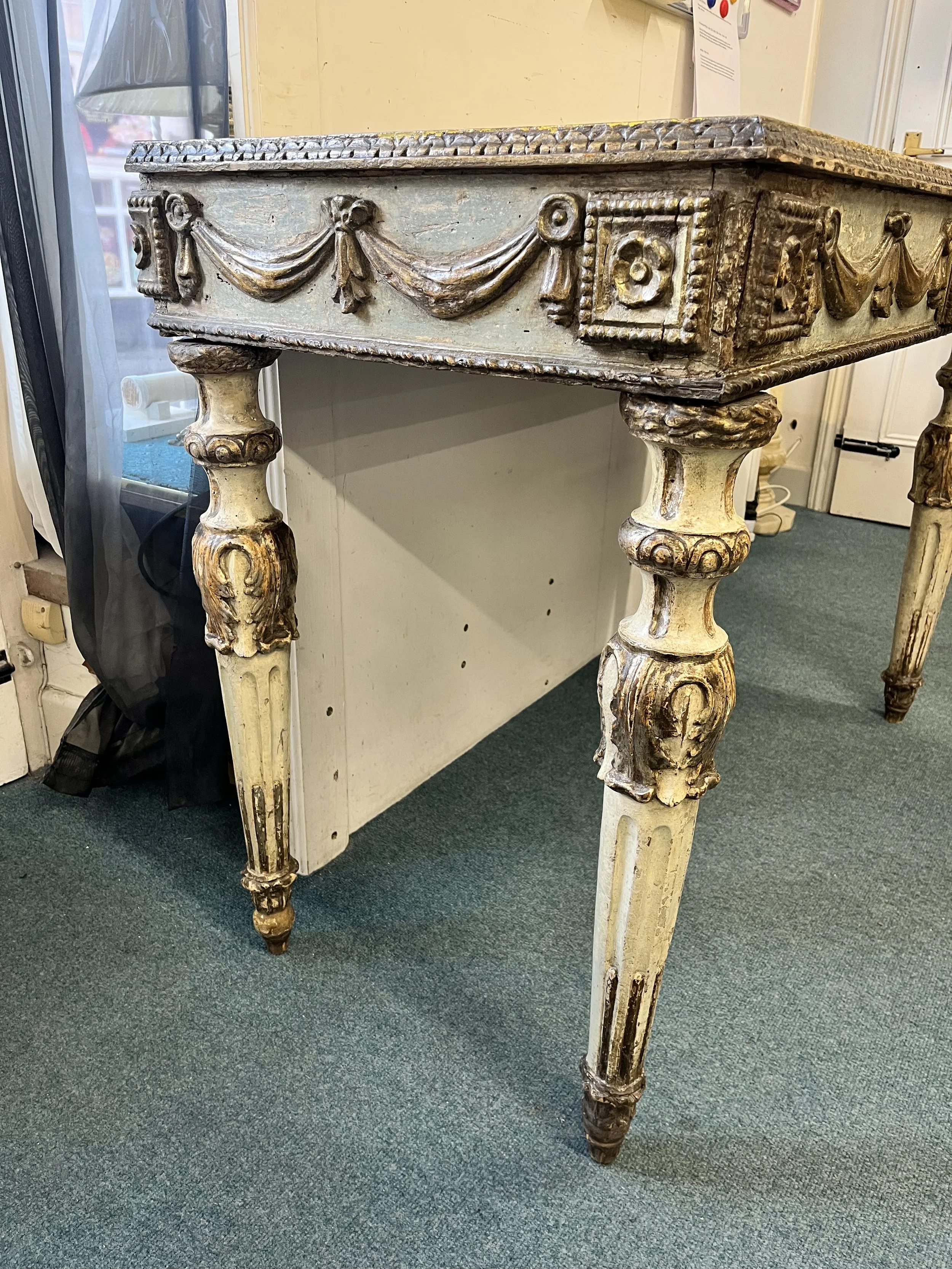 An antique wooden table with ornate carved details, painted in a distressed cream color, standing on a green carpeted floor.