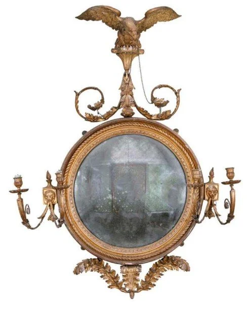 An ornate antique mirror with a round glass, surrounded by a gold frame, adorned with candle holders on each side, a decorative eagle with outstretched wings on top, and intricate gold detailing.