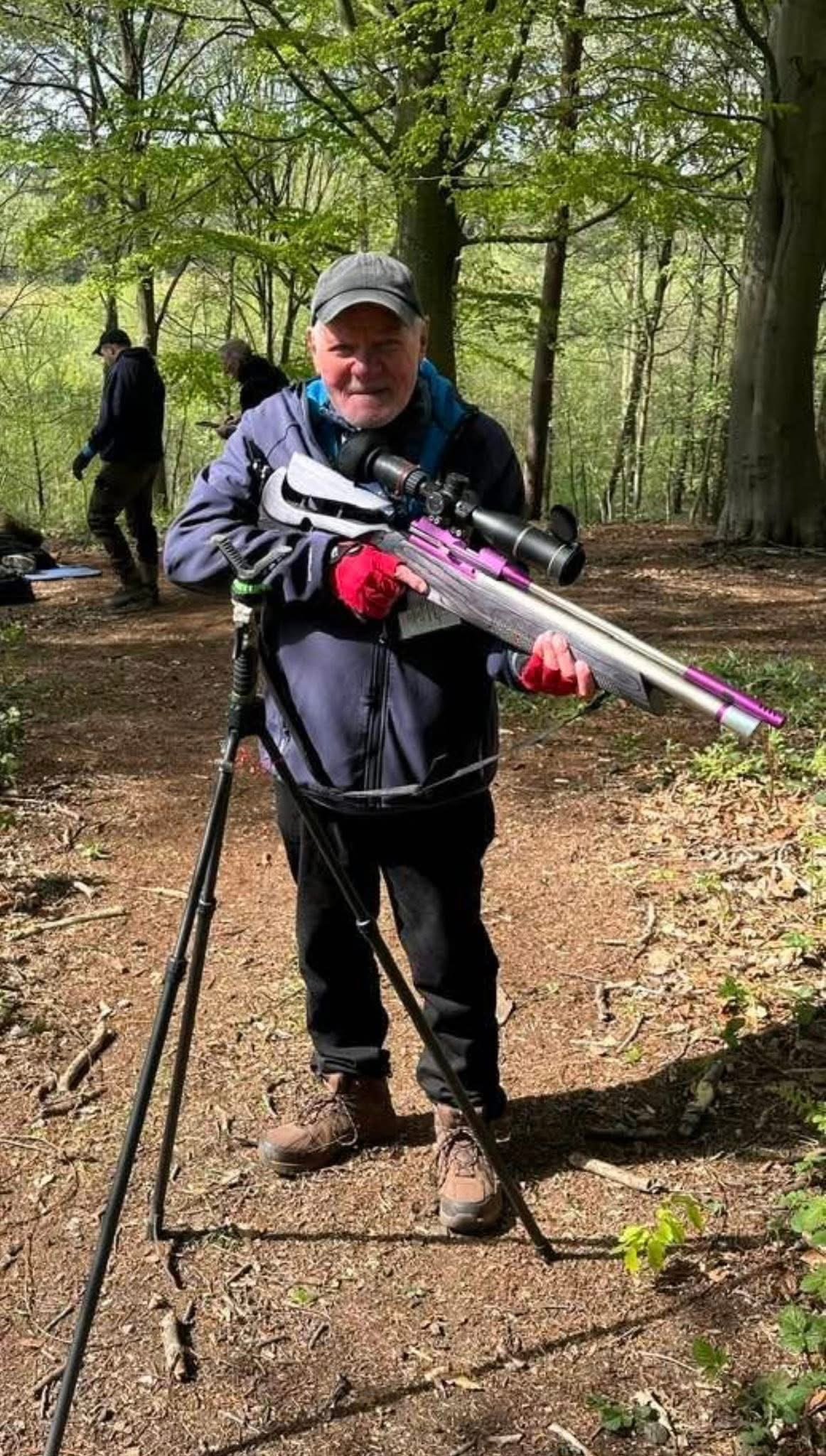 Older man in outdoor clothing and a cap holding a rifle with a scope, standing on a forest trail, with two people in the background in a wooded area.