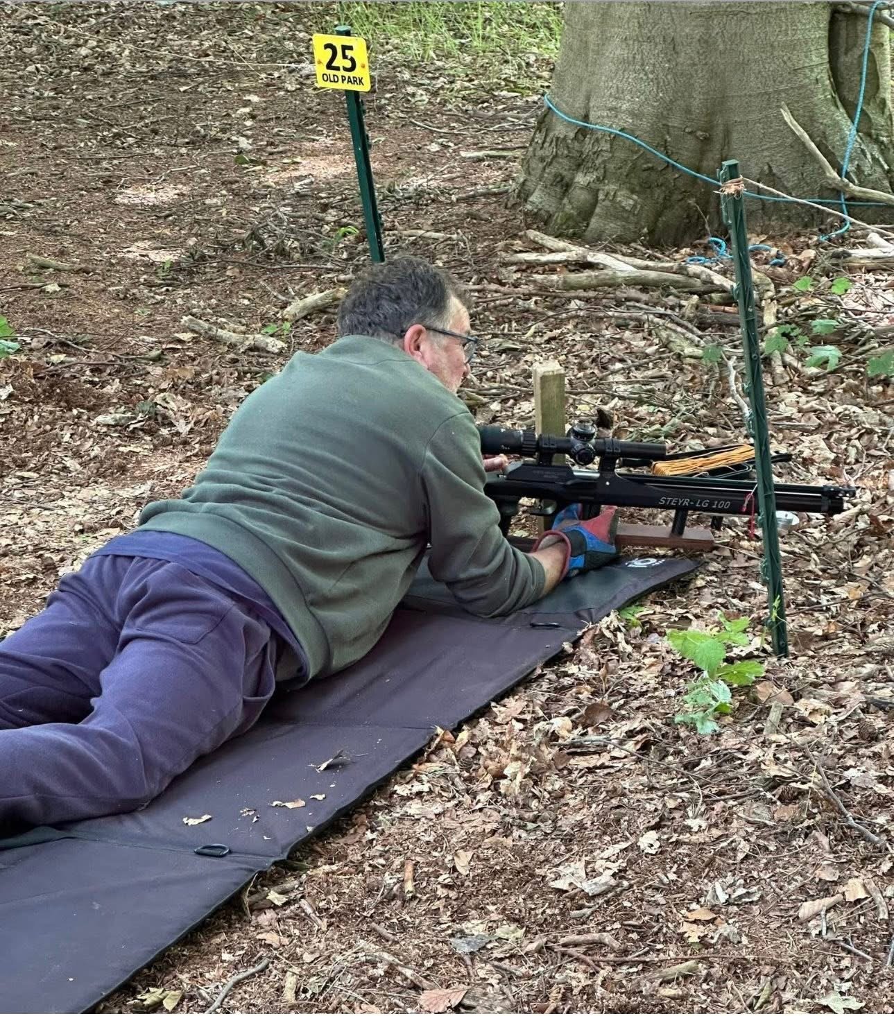 A man lying on his stomach on a black mat in the woods, aiming with a rifle through a scope. There is a yellow sign in the background that says '25 Old Park'.