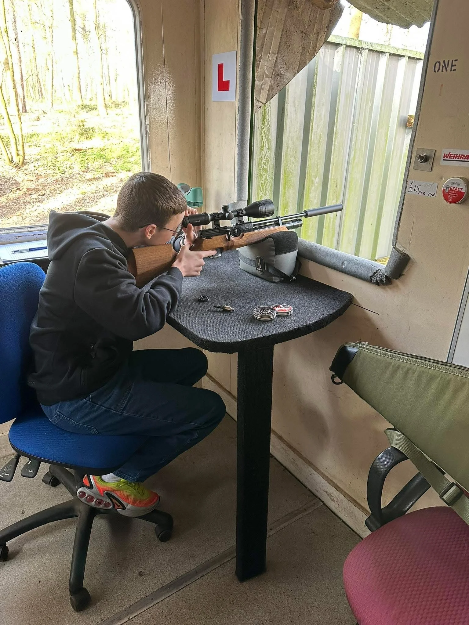 A young boy in a black hoodie and colorful sneakers is sitting on an office chair, aiming a wooden rifle with a scope through a window at a shooting range.