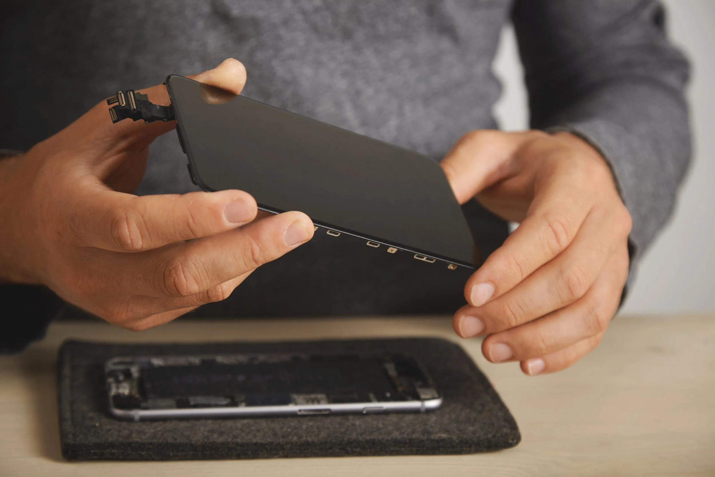 Person holding a black smartphone with its screen facing inward, appearing to work on it, with a disassembled phone or parts on the table below.