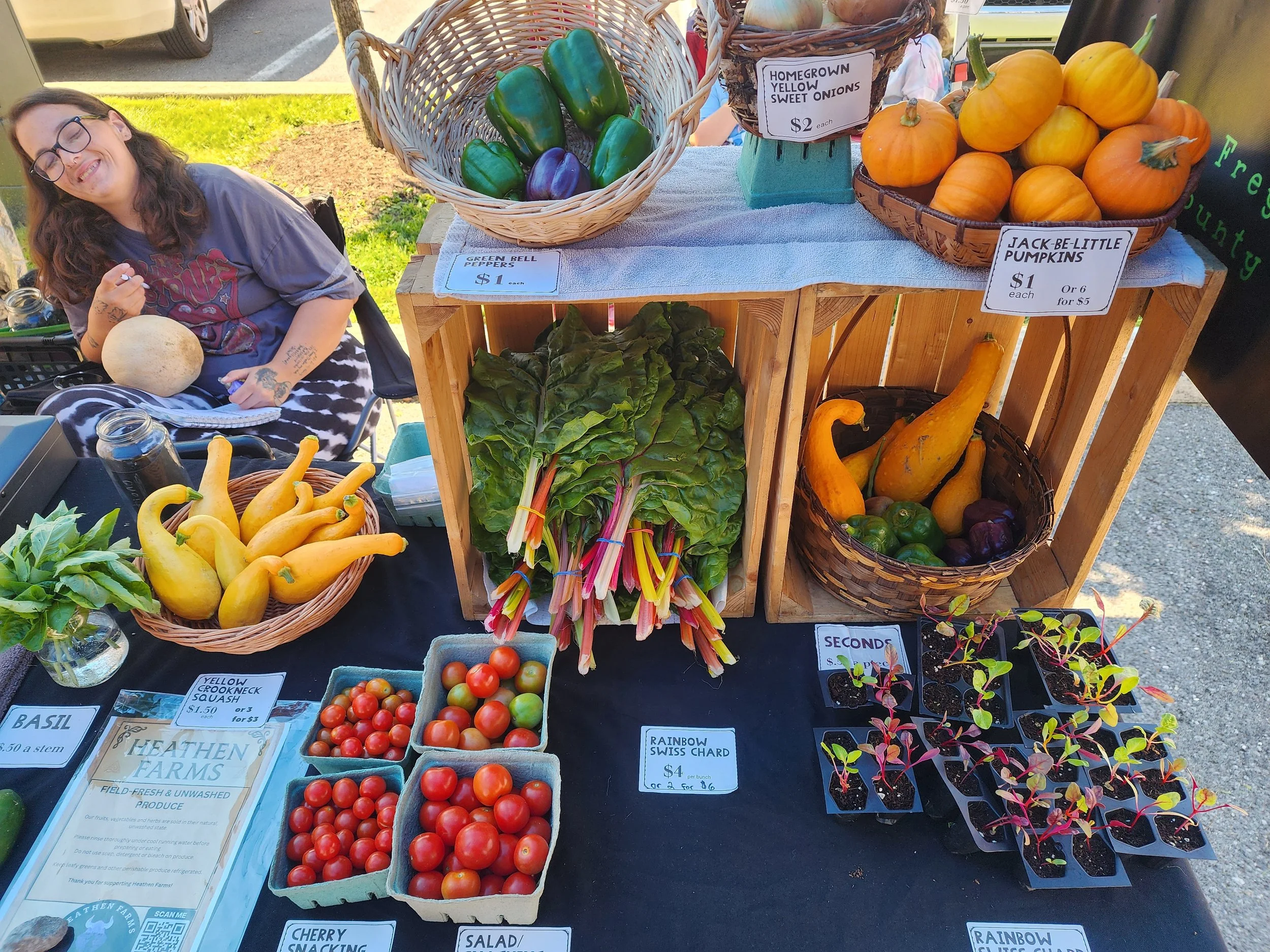 At a farmer's market stall, there are baskets with fresh produce, including yellow crookneck squash, green bell peppers, heirloom tomatoes, and jack-be-little pumpkins. Signs indicate prices, and a woman with glasses and tattoos is smiling and sitting near the stall, holding a squash.