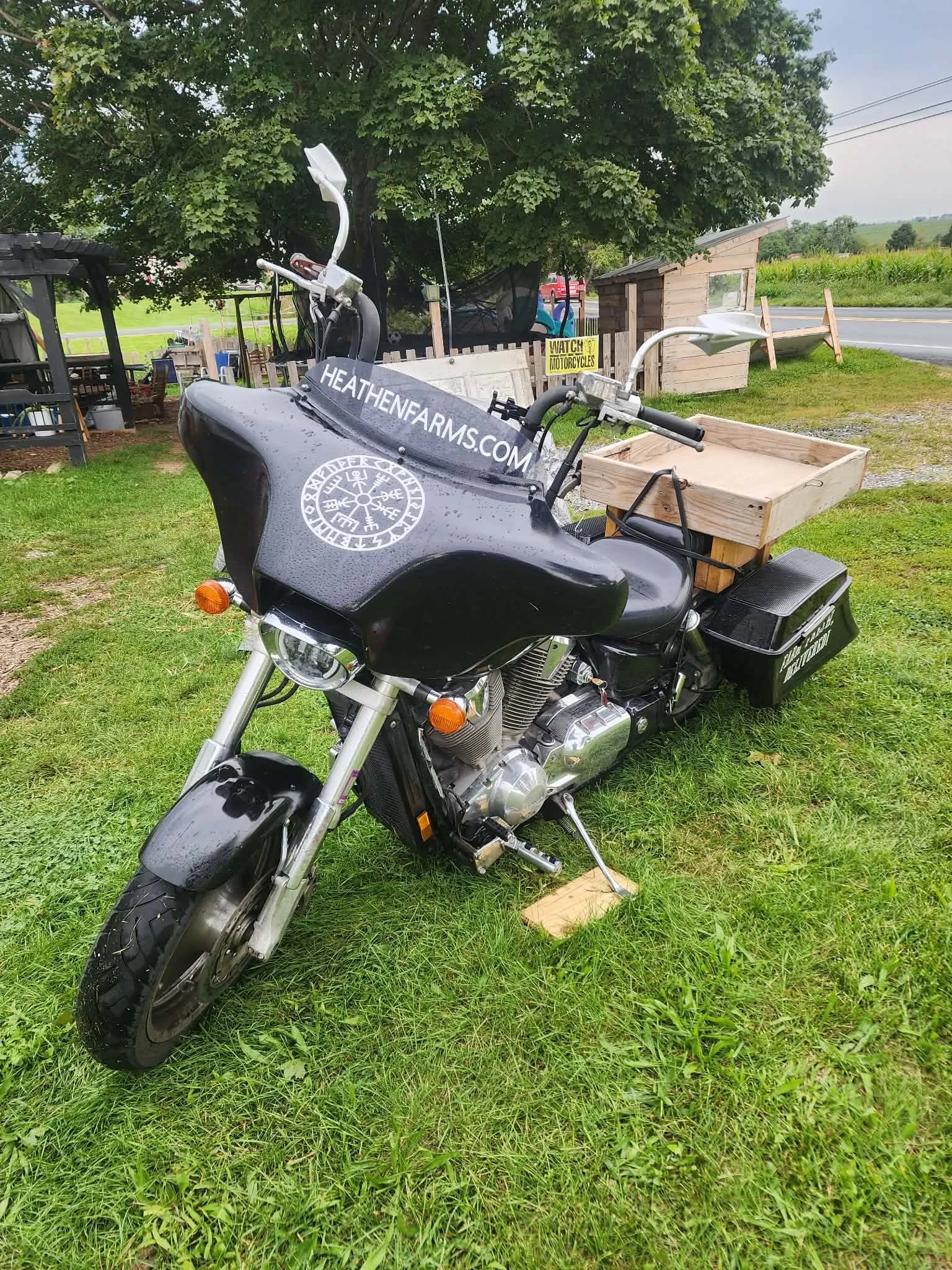 A black motorcycle with a wooden crate attached to the back, parked on a grassy area near a rural road, with trees and a small shed in the background.