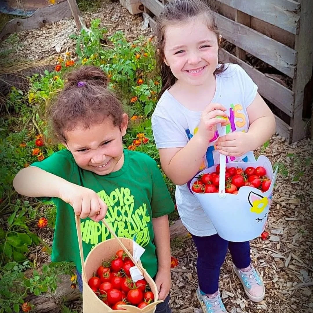 Two girls smiling and holding baskets of cherry tomatoes in a garden filled with plants and flowers.