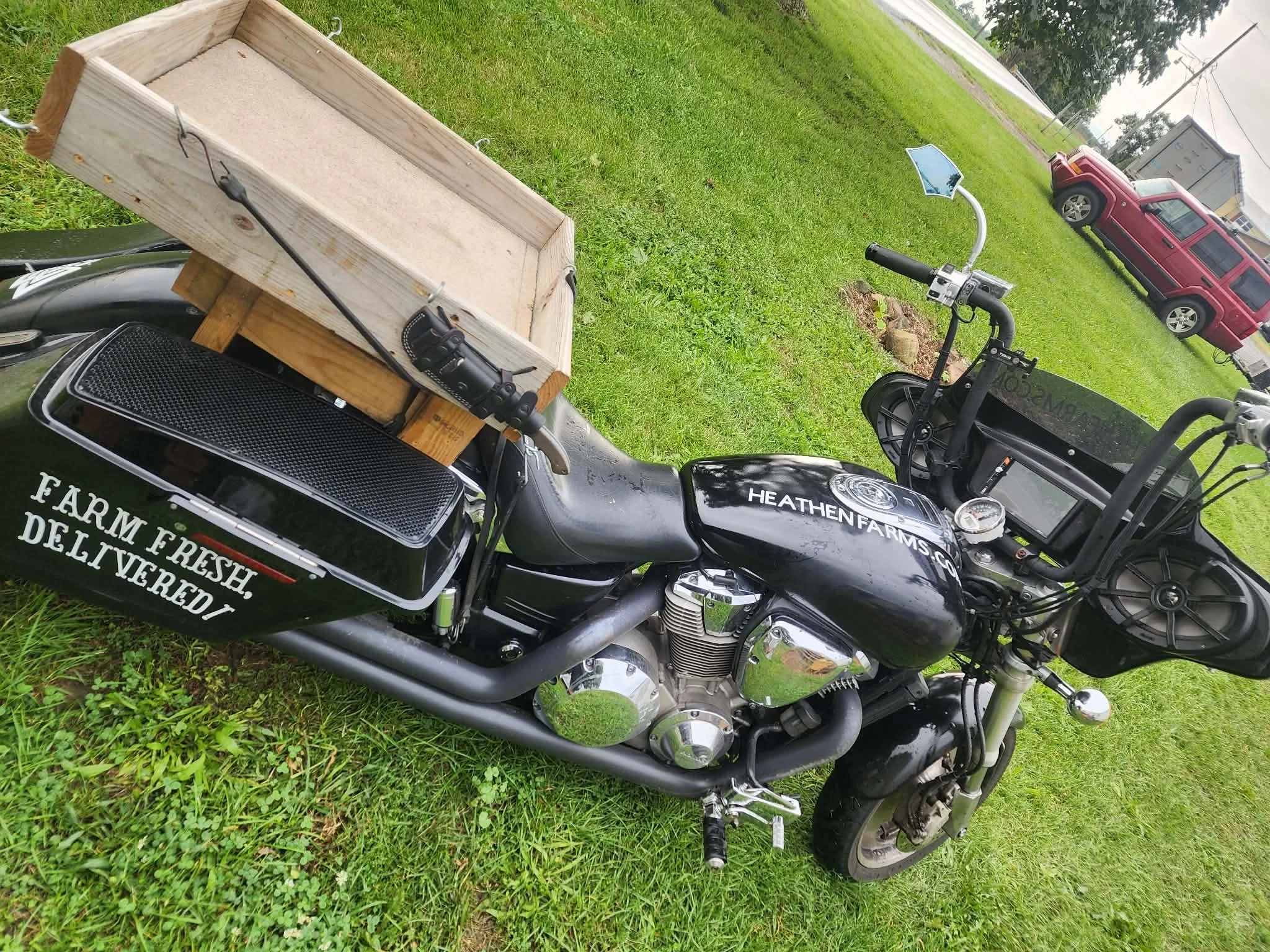 A black motorcycle with a wooden delivery box attached to the back, parked on a grassy area near a road, with a red vehicle and some trees in the background.