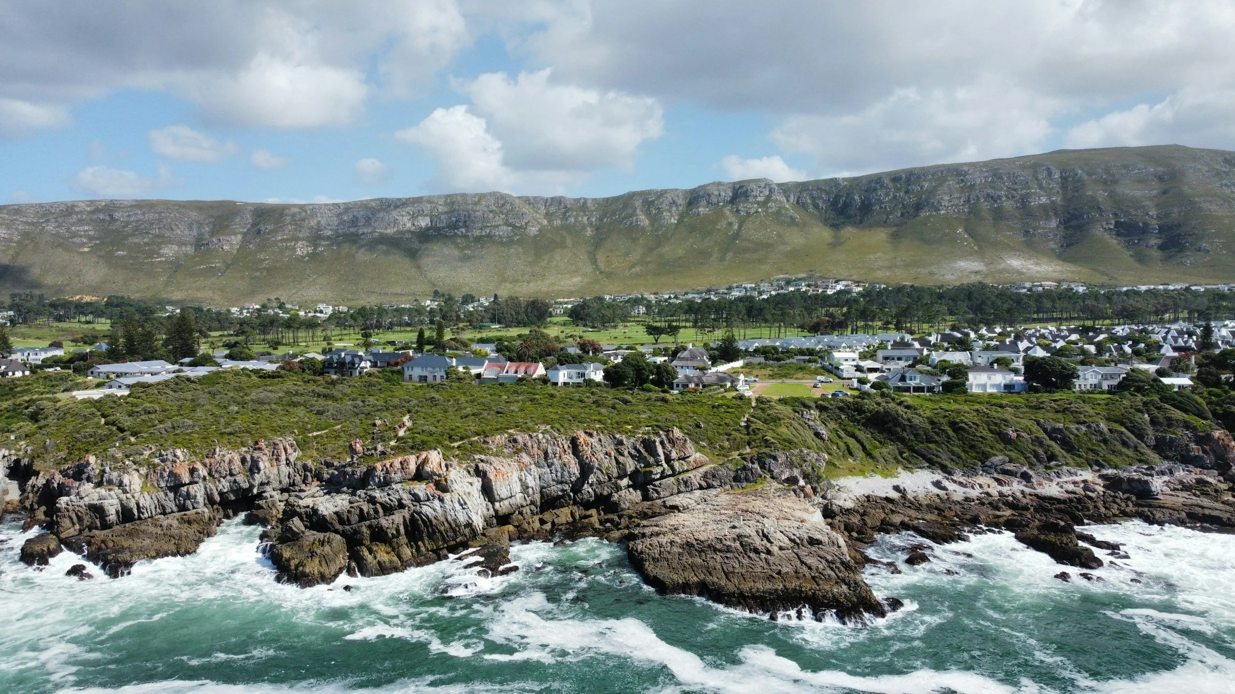 Coastal town with houses at the base of green rolling hills and rocky cliffs, ocean waves crashing along the shore under partly cloudy sky.