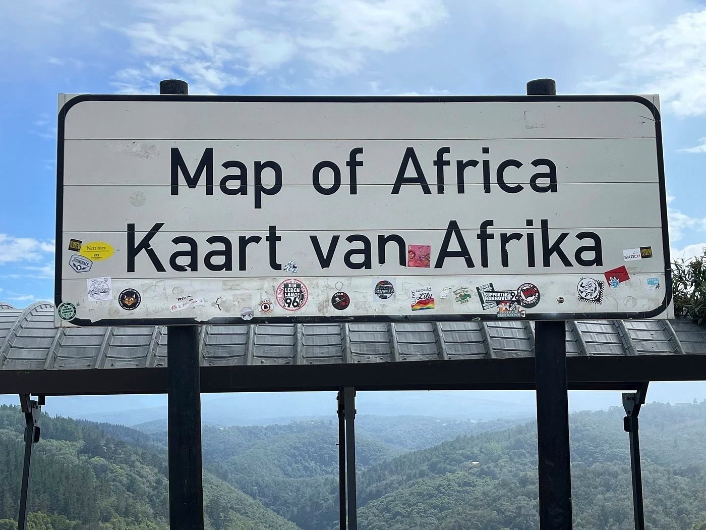 Sign that reads 'Map of Africa' in English and Dutch, decorated with stickers, against a mountain landscape and blue sky with clouds.
