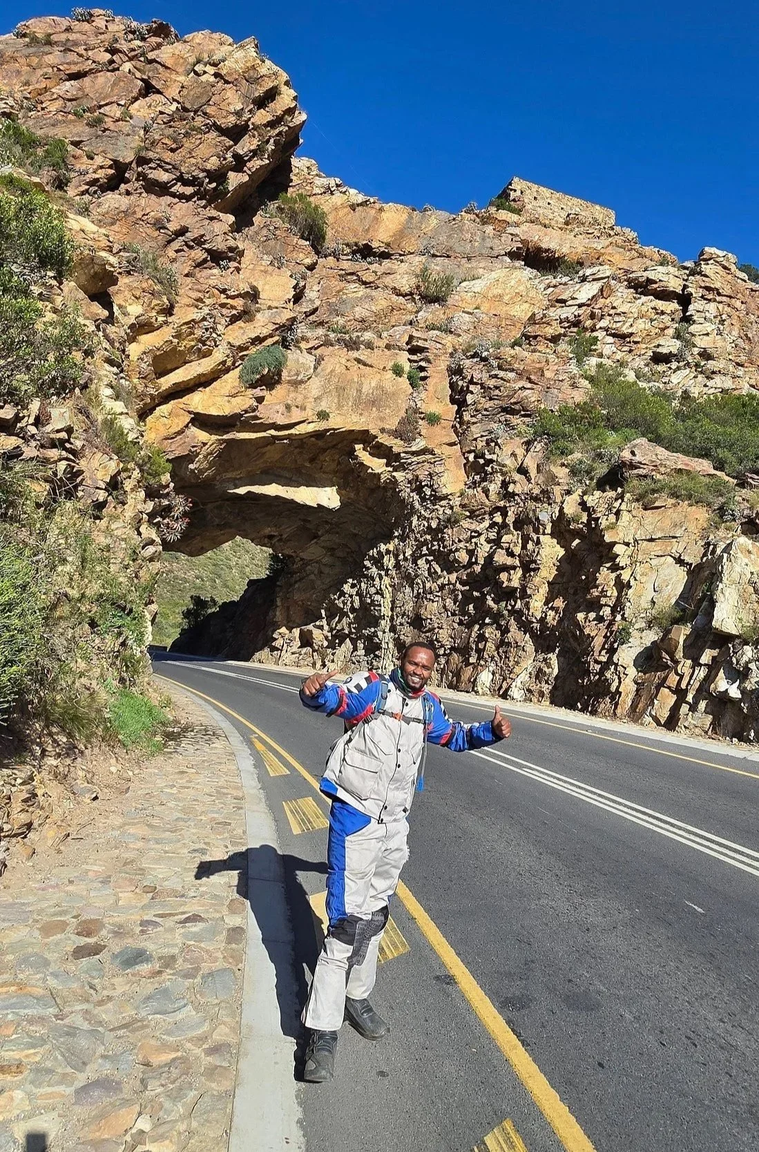 A man standing on the side of a road near a rock formation with a natural arch and a tunnel. He is smiling and giving two thumbs up, wearing a motocross outfit. The sky is clear and blue.