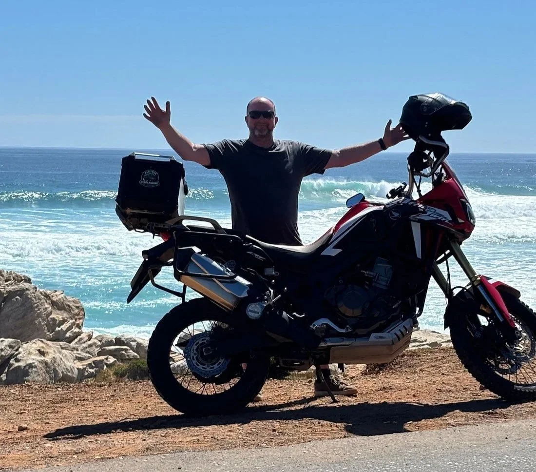 A man standing next to a black motorcycle with a red front, on a dirt road near the ocean, with waves and blue sky in the background, holding a black helmet and wearing sunglasses. Misty Cliffs Scarboughrough Cape Town Southern Suburbs South Africa 