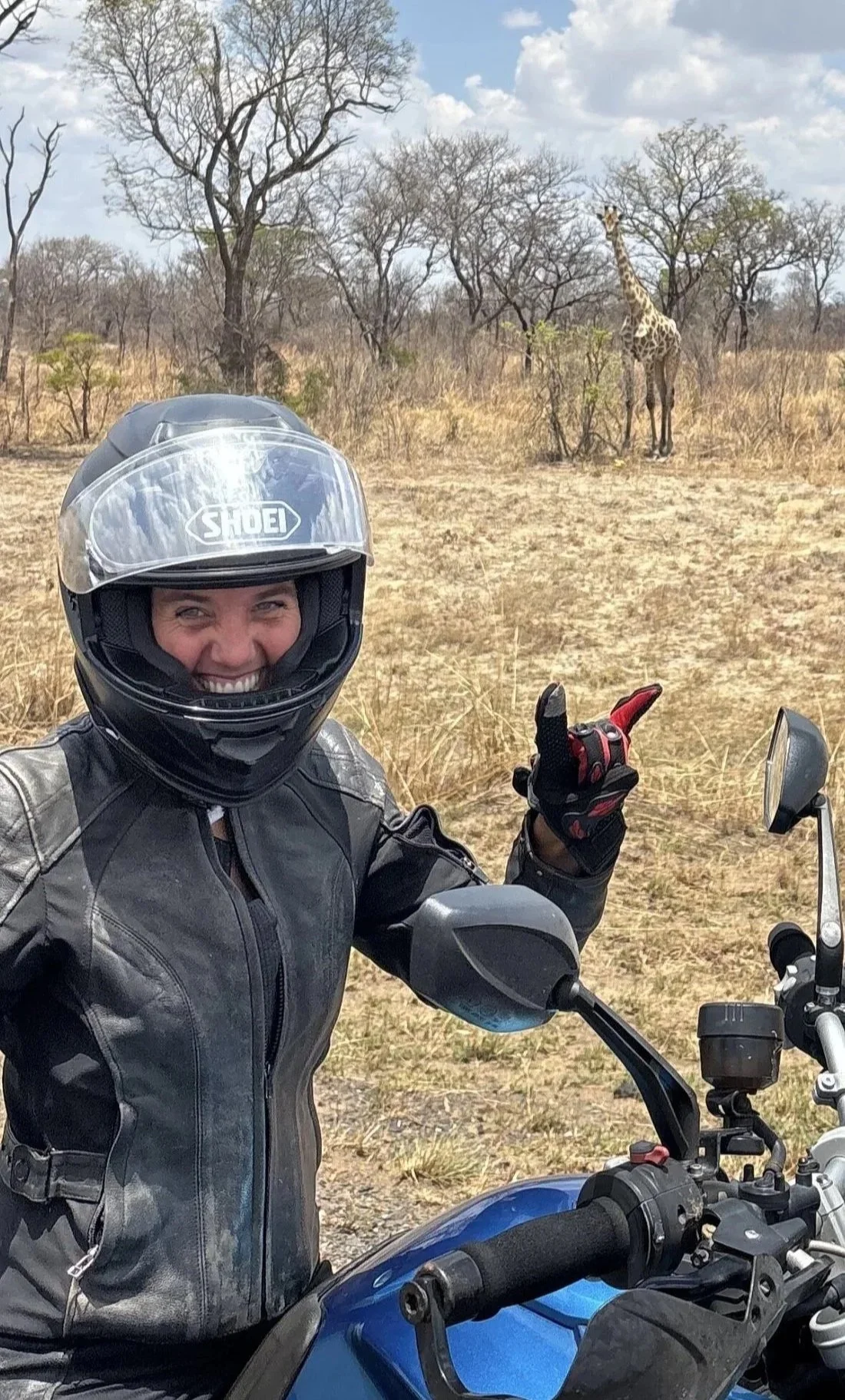 A woman in motorcycle gear taking a selfie on her motorcycle in a dry savanna with a giraffe in the background