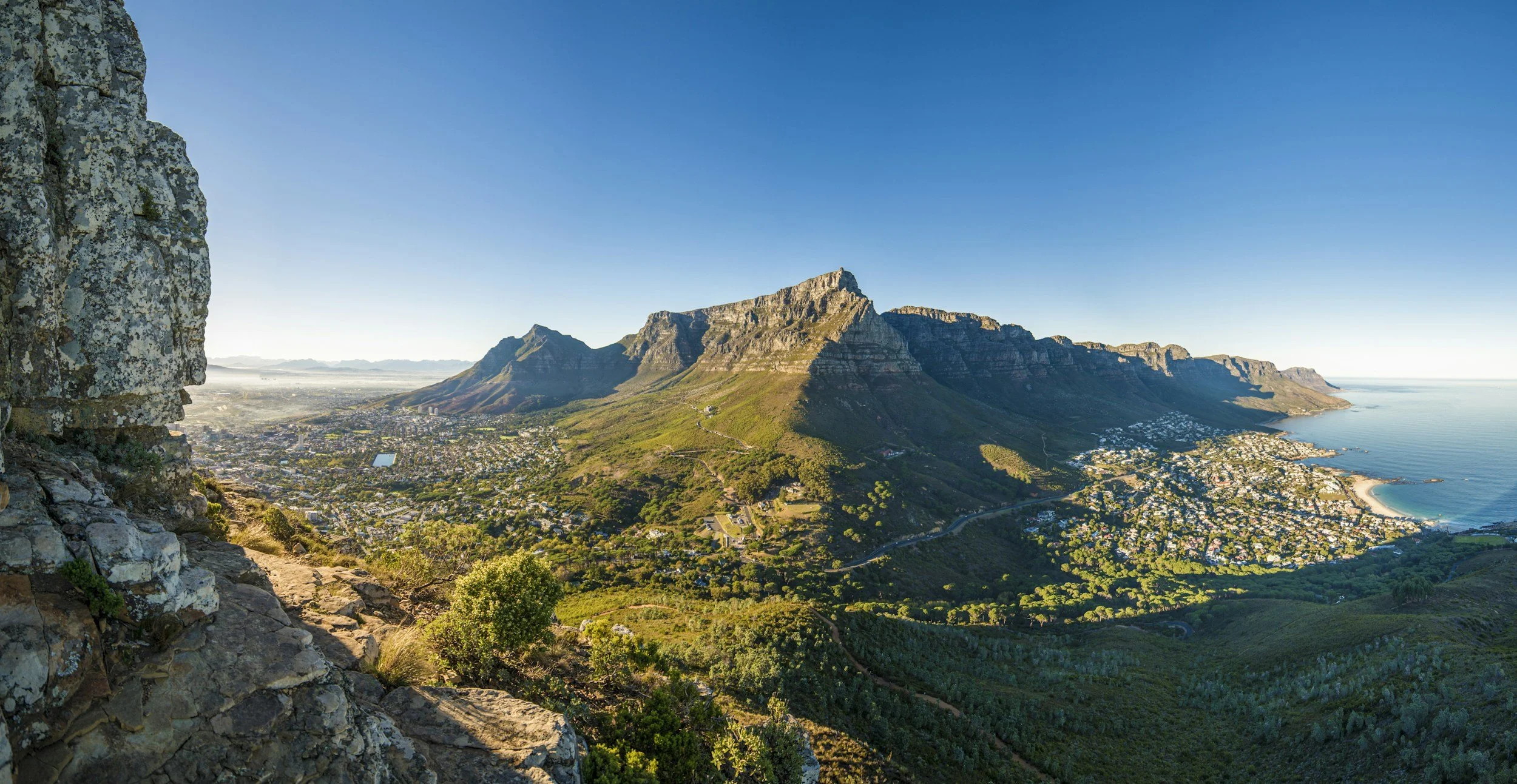 A scenic view of a coastal city with a mountain range in the background, seen from a high vantage point.