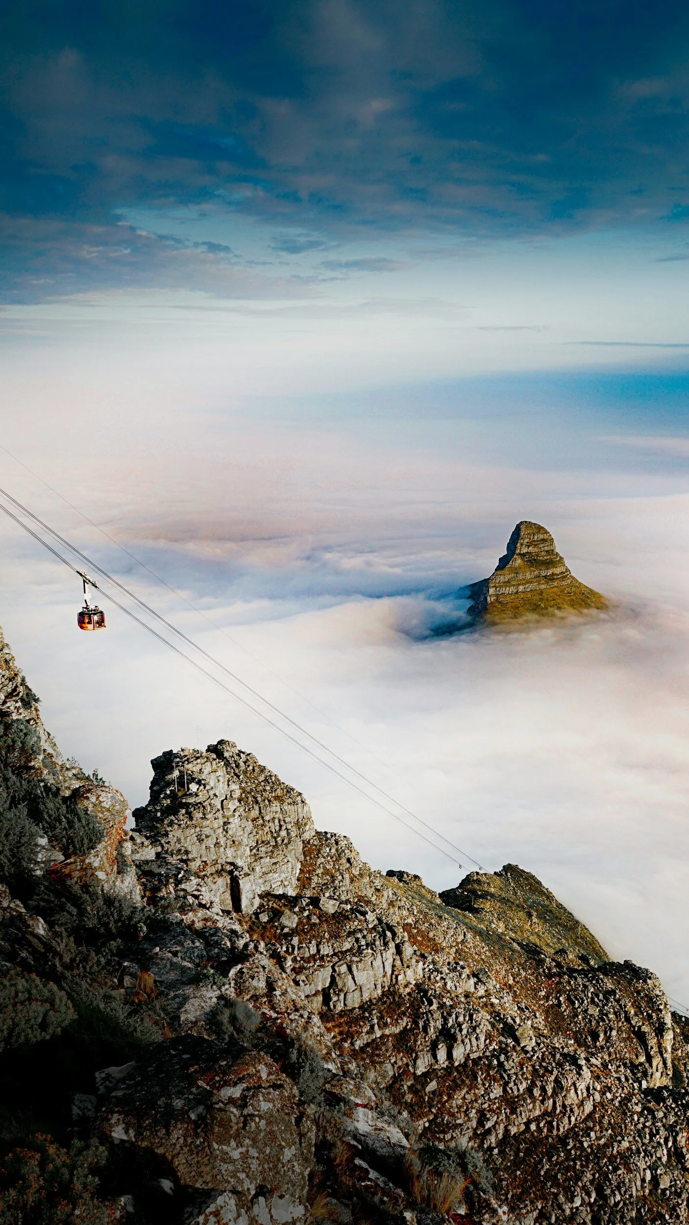 A cable car ascending above a mountain landscape shrouded in clouds, with rocky peaks and a mountain peak in the distance.